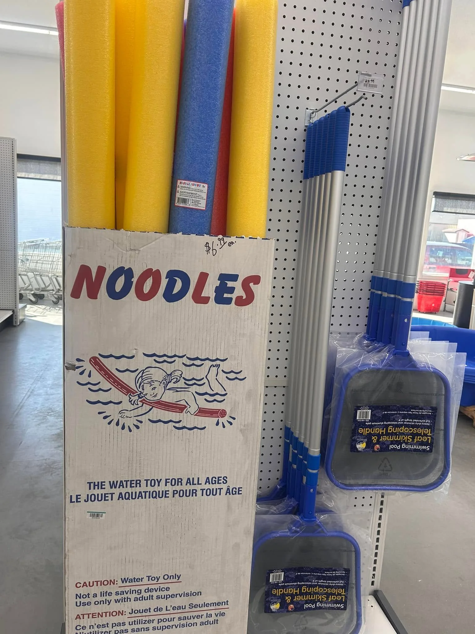 Large cardboard box containing colorful water toys, labeled for use by children of all ages, with a surfboard-like design on the front. To the right, blue and silver pool skimmers are displayed hanging on a pegboard.