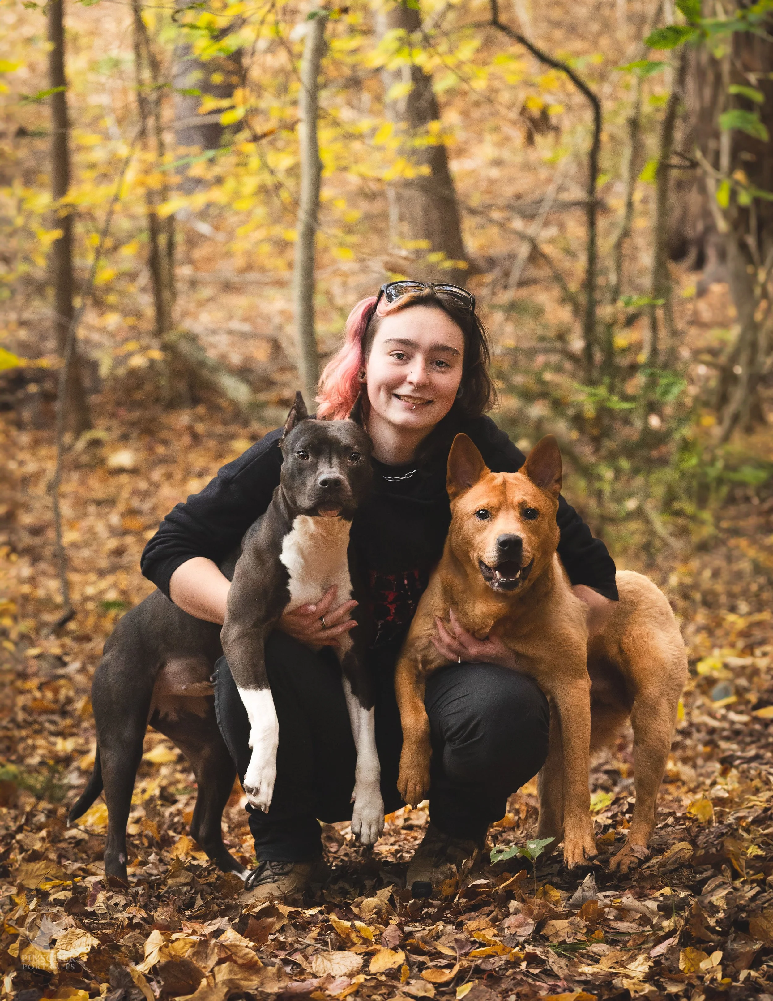 A non-binary person with pink and brown hair crouching in a forest with fallen autumn leaves, smiling and holding two dogs, one black and white and one brown.