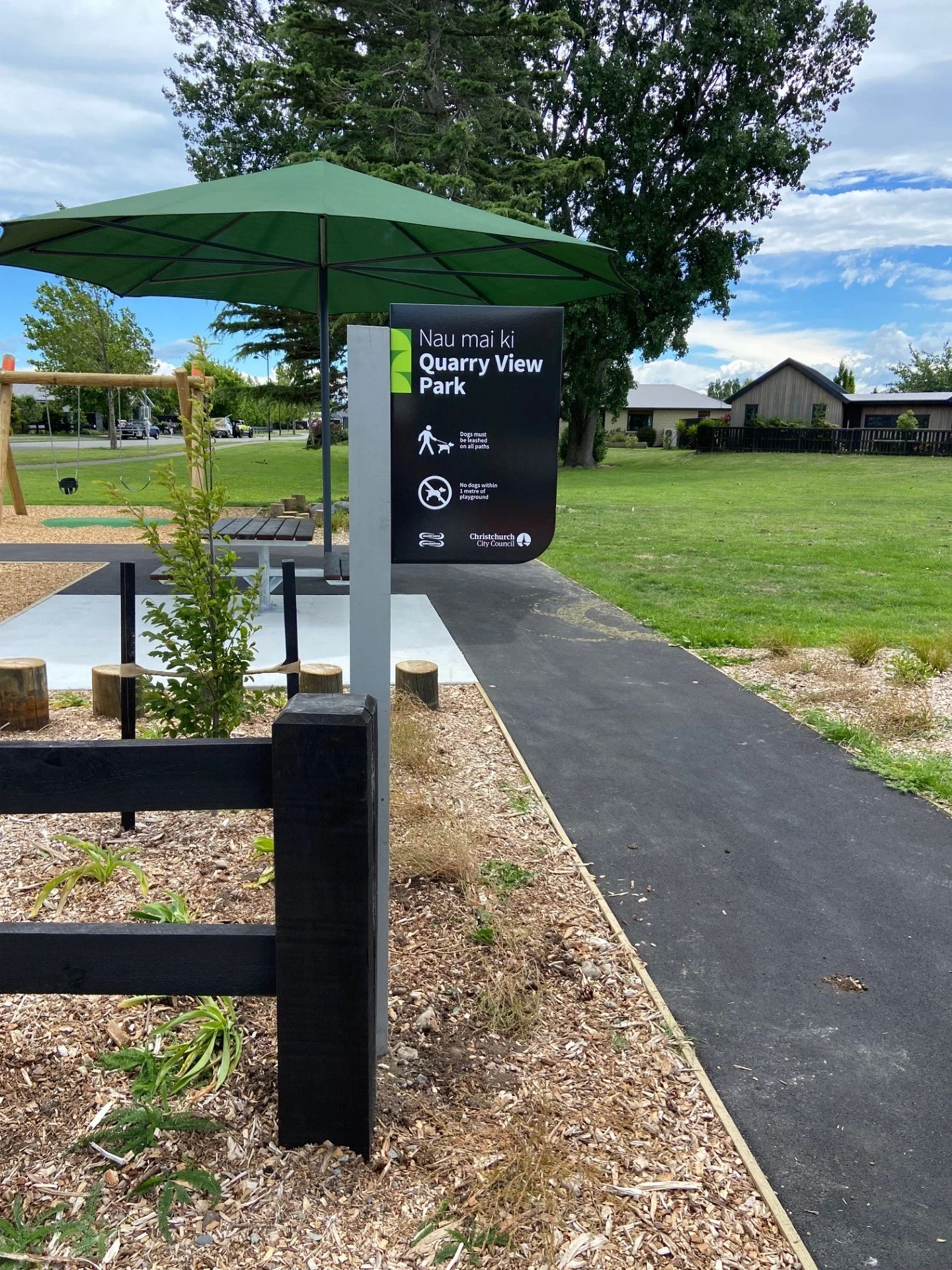 Park entrance sign for Nau mai ki Quarry View Park with rules for dogs, under a green umbrella, with a paved walking path, grass, trees, and residential buildings in the background.