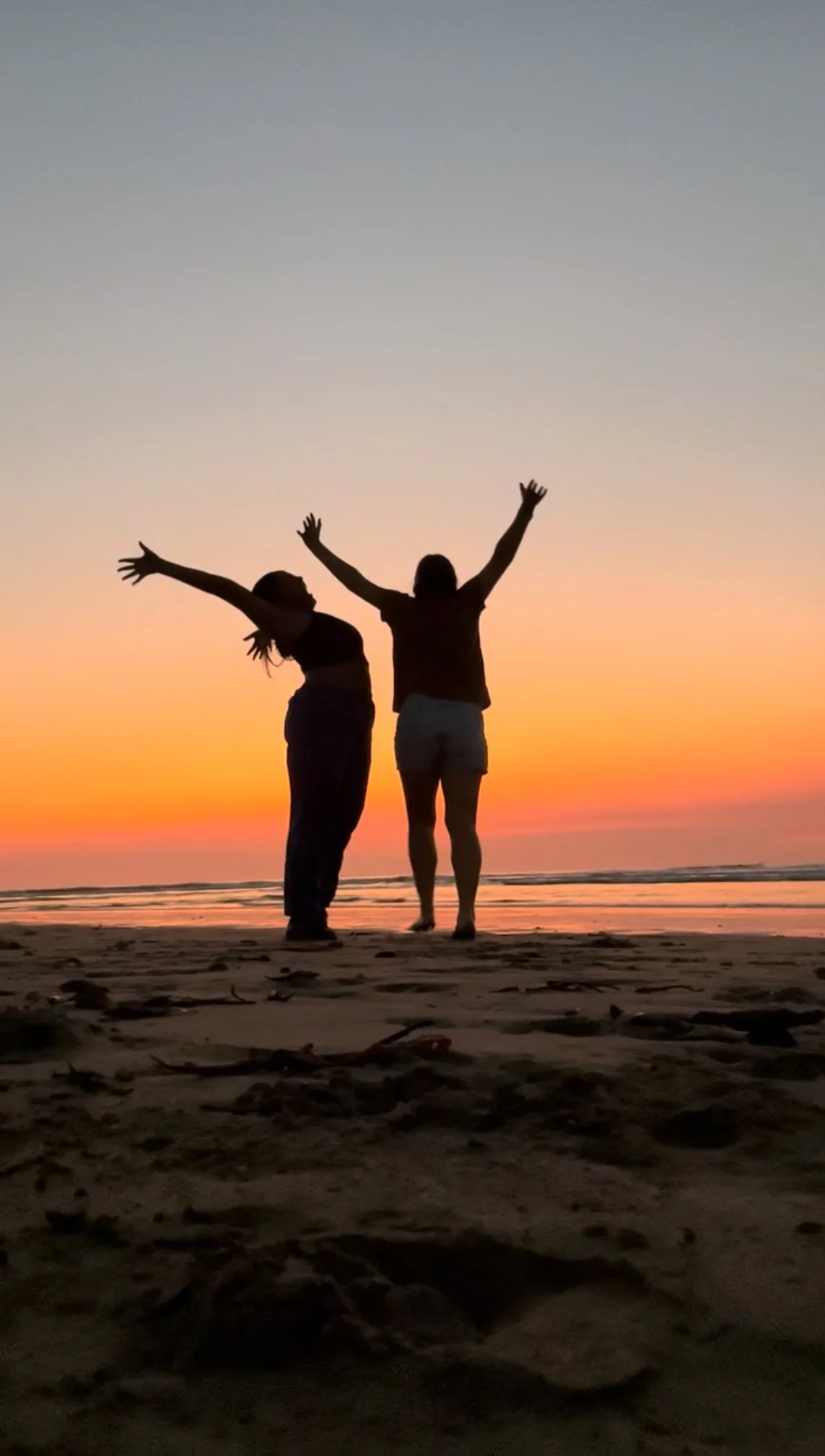 Silhouette of two women standing on the beach during sunset, with arms raised and joyfully posing near the shoreline.