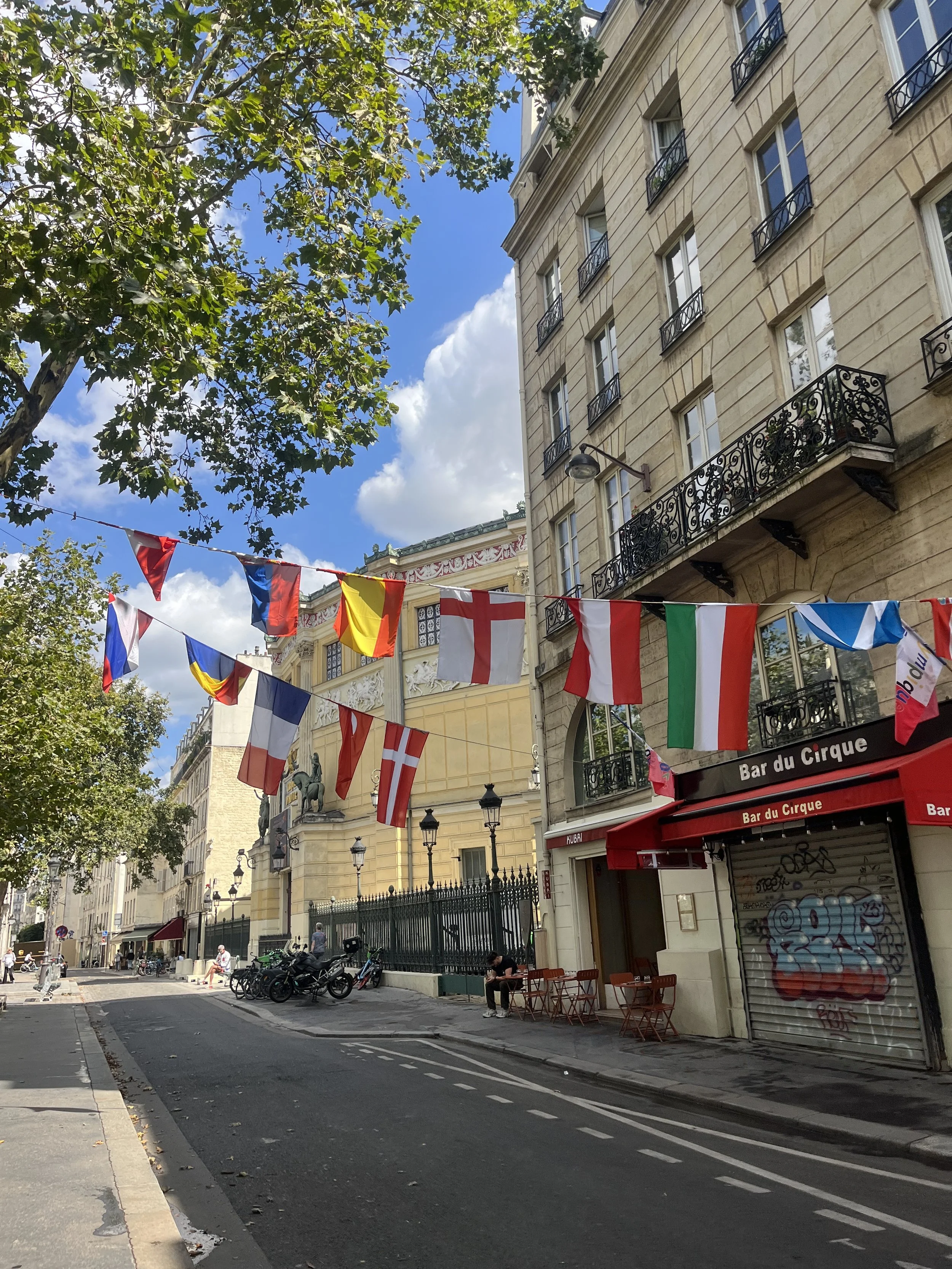 Paris street scene with colorful flags strung across the sky, classic European architecture with balconies, trees, and a street cafe with graffiti on the shutter.