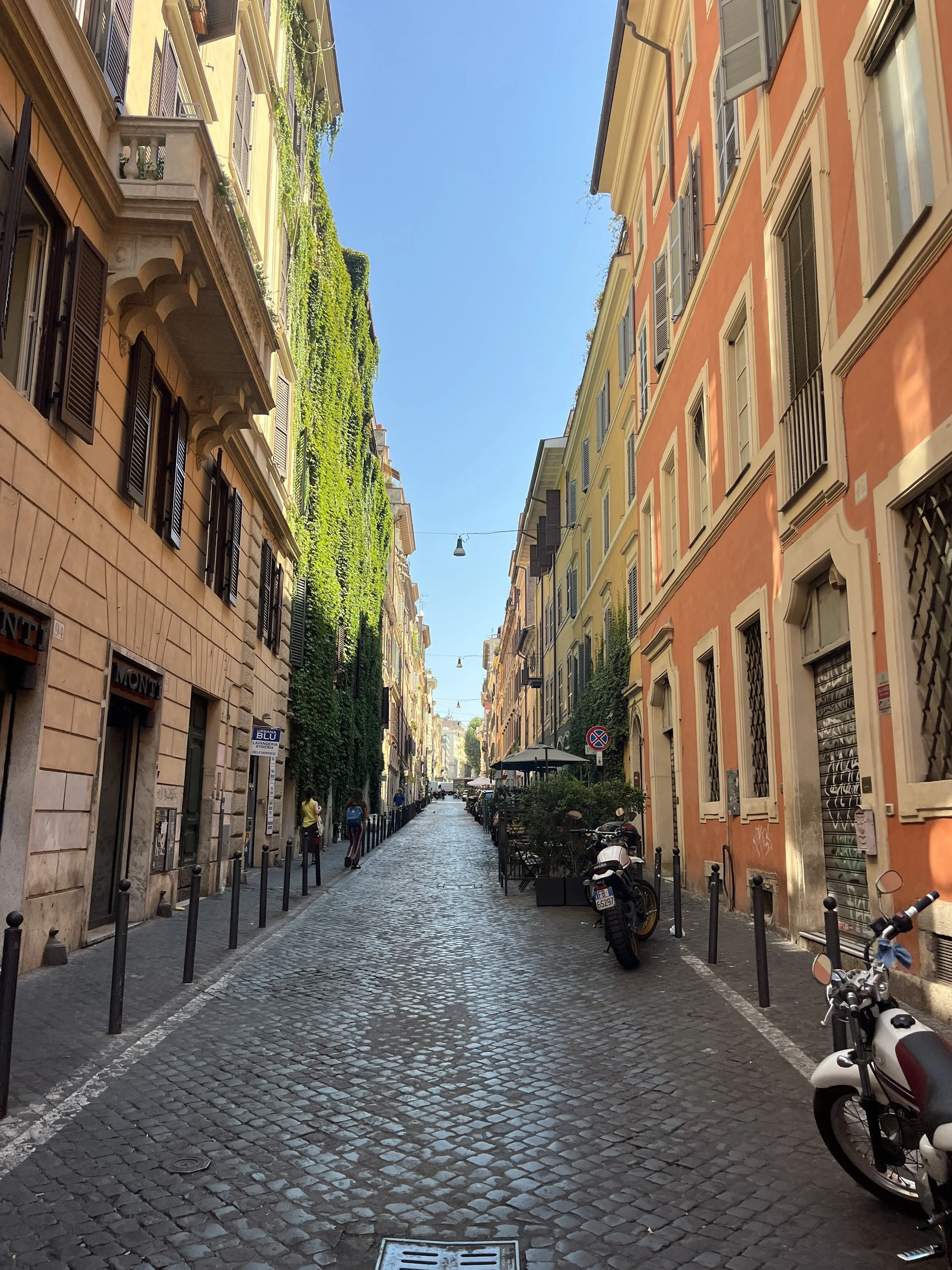 A narrow cobblestone street lined with colorful European-style buildings, some with green ivy, under a clear blue sky. There are parked motorcycles and a few pedestrians.
