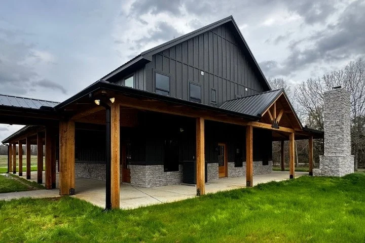 Dark gray timber frame home with wrap-around covered porch, stone chimney, and metal roof