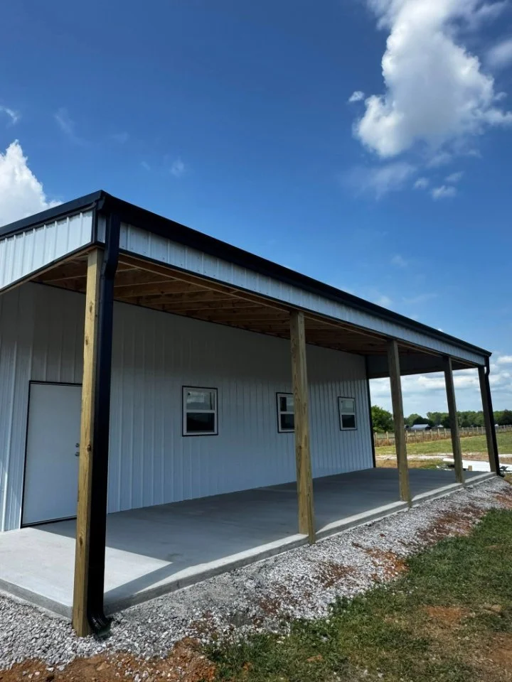 Exterior of a new metal building under construction with a raised porch, wooden support beams, and a gravel area in front, set against a bright blue sky with clouds.