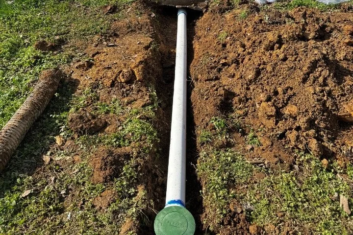 White PVC underground drain pipe laid in an open dirt trench with a green pop-up drain cap at the end
