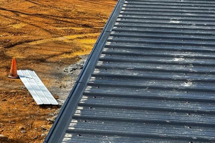 Close-up of a seamless gutter installed along the edge of a dark corrugated metal roof on a construction site with an orange traffic cone on the ground