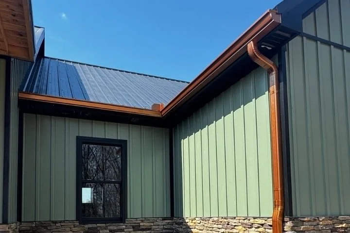 Copper seamless gutter and downspout running along the corner of a green metal home with stone veneer and a black window