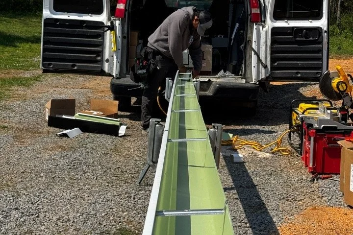 Person fabricating a custom green seamless gutter section at the back of a work van on a job site