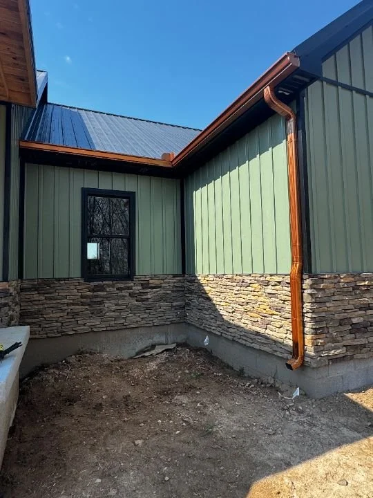 Exterior corner of a house with green metal siding, stone veneer at the bottom, and copper gutter and downspout against a blue sky.
