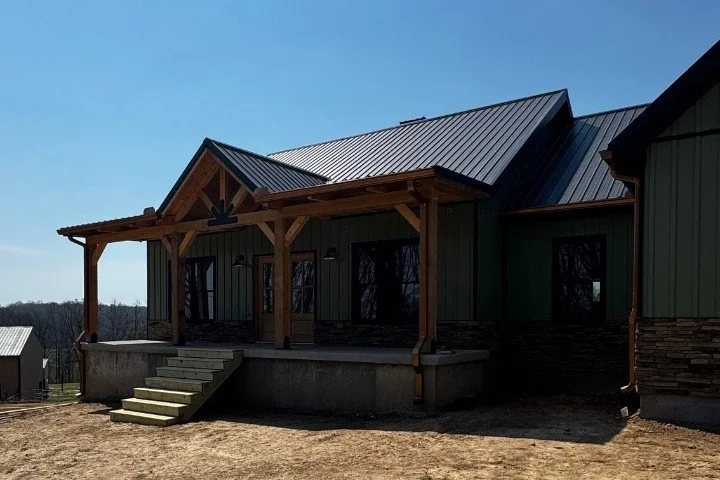Front view of a new house with a metal roof, wooden porch, and steps leading up to the entrance, set in a rural area with a clear blue sky.