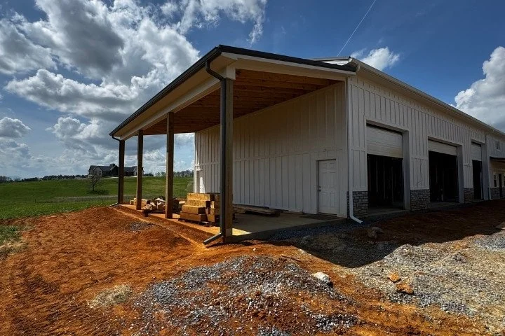 White metal shop building with black seamless gutters, three garage bays, a covered timber porch, and stacked lumber on a rural property