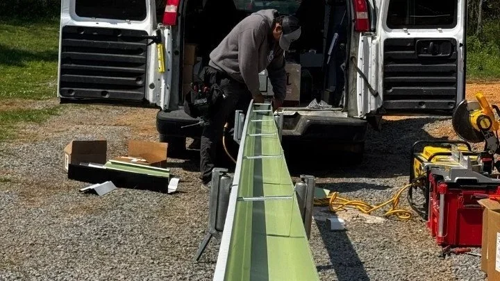 A worker inspects or installs a long, green, metal structure, possibly a part of equipment or infrastructure, with the back of a white van in the background and tools and supplies nearby.