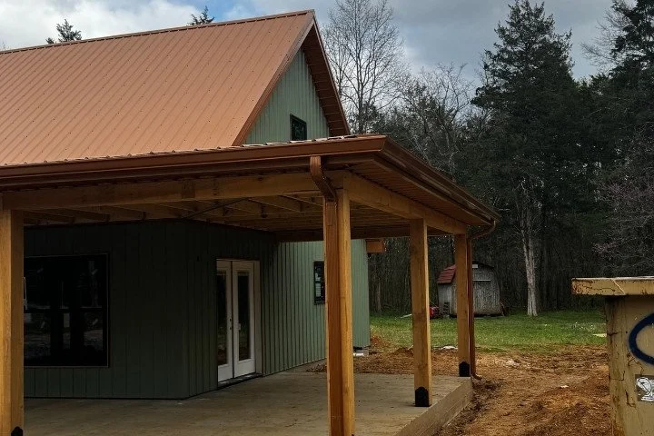 Green house with a brown metal roof and a covered porch with wooden posts, surrounded by trees and a grassy yard.