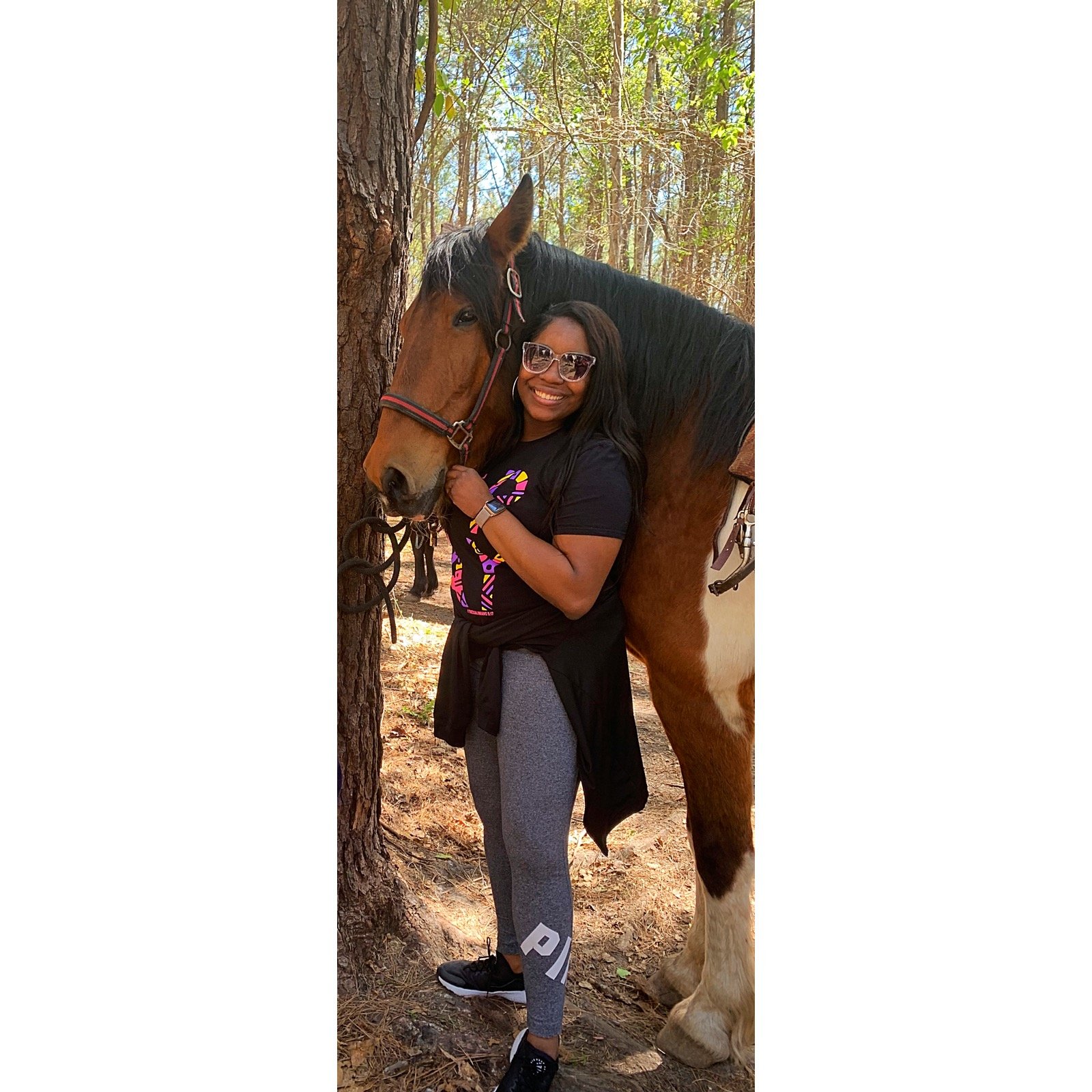 A woman standing next to a large brown and white horse in a wooded area, smiling and holding the horse's bridle.