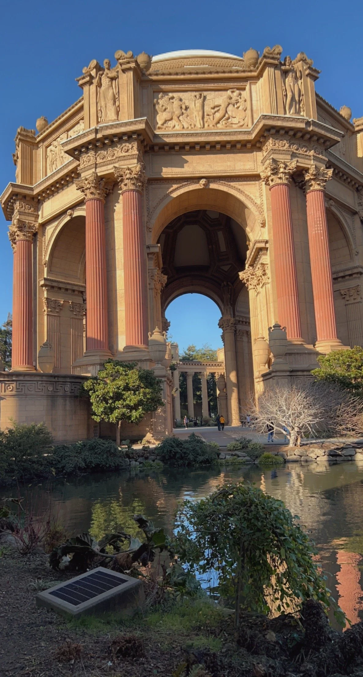 Large historic monument with Corinthian columns, ornate carvings, and a fountain in the foreground, under a clear blue sky.