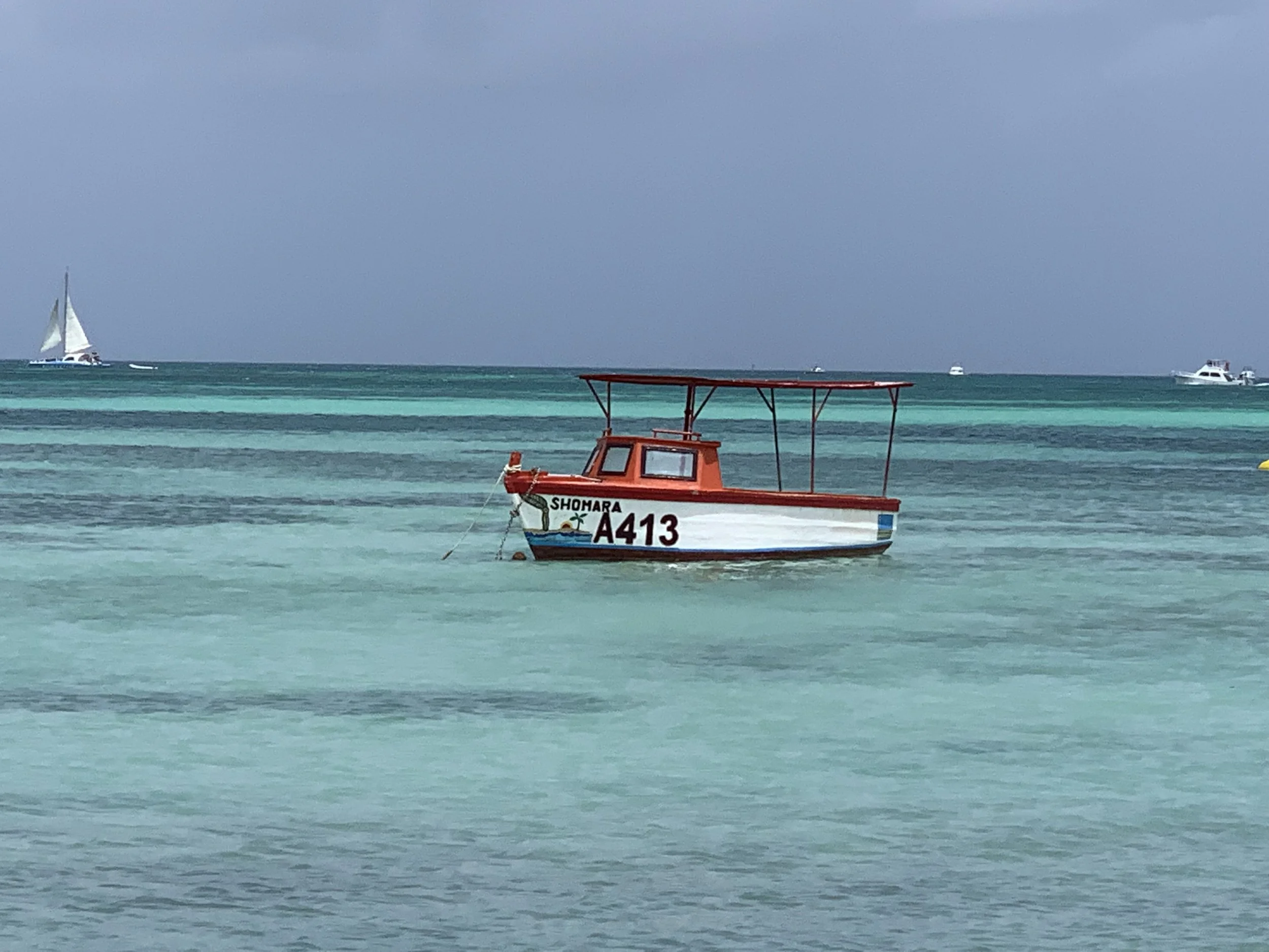 A small red and white boat named Shomara A413 floating on turquoise water, with sailboats in the background under a blue sky.