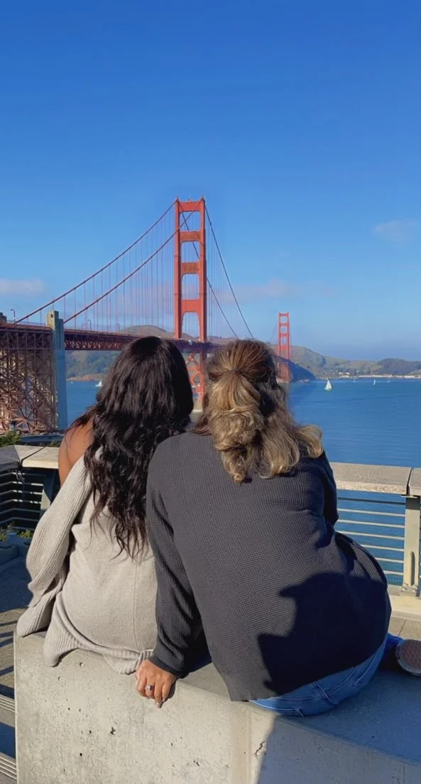 Two women sitting on a concrete ledge, overlooking the Golden Gate Bridge in San Francisco, with sailboats in the water and a clear blue sky.