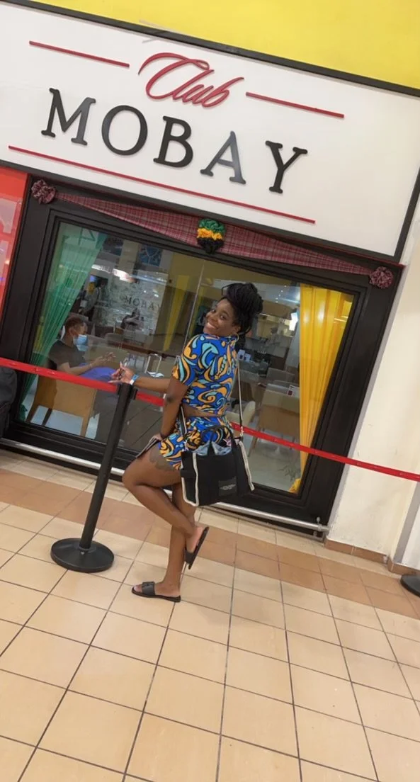 A woman stands in front of Club Mobay, a luggage service at the airport, posing playfully with one foot raised and smiling.