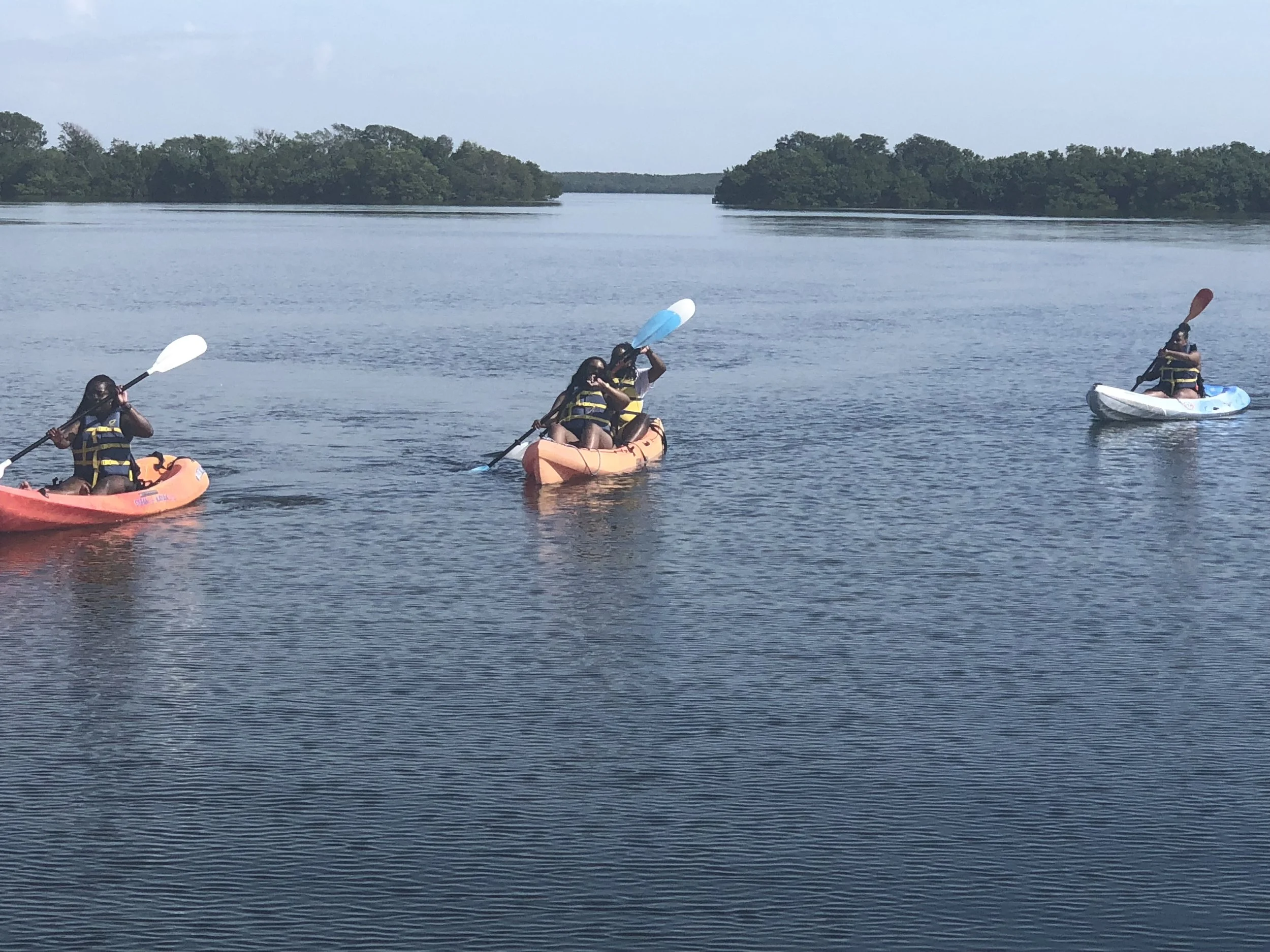 Three children kayaking on a calm body of water with trees in the background.