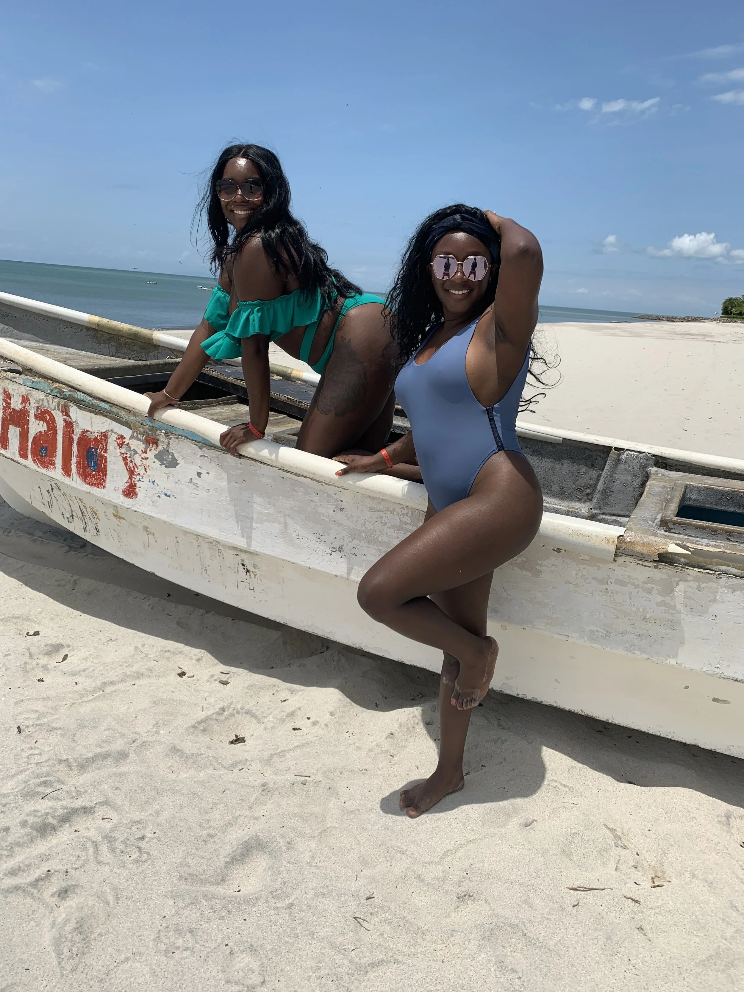 Two women on a sandy beach, posing with a small boat. One woman is kneeling on the boat, wearing a green ruffled bikini top and glasses. The other woman is standing with one leg raised, wearing a blue one-piece swimsuit and sunglasses. The ocean and 