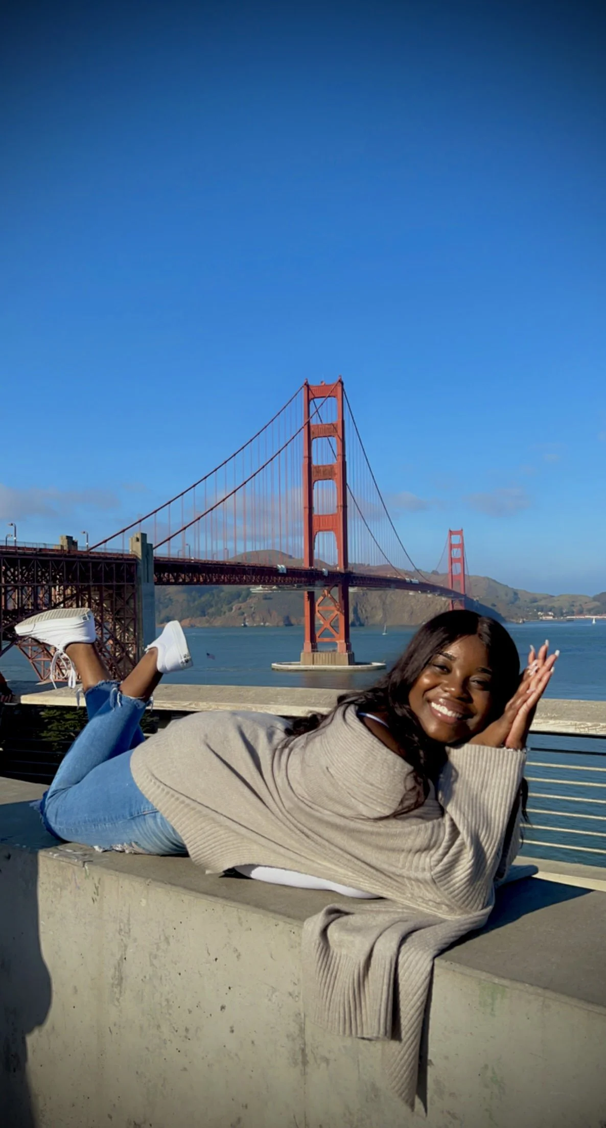 A smiling woman lying on a concrete ledge near the Golden Gate Bridge in San Francisco, California, with water and hills in the background.