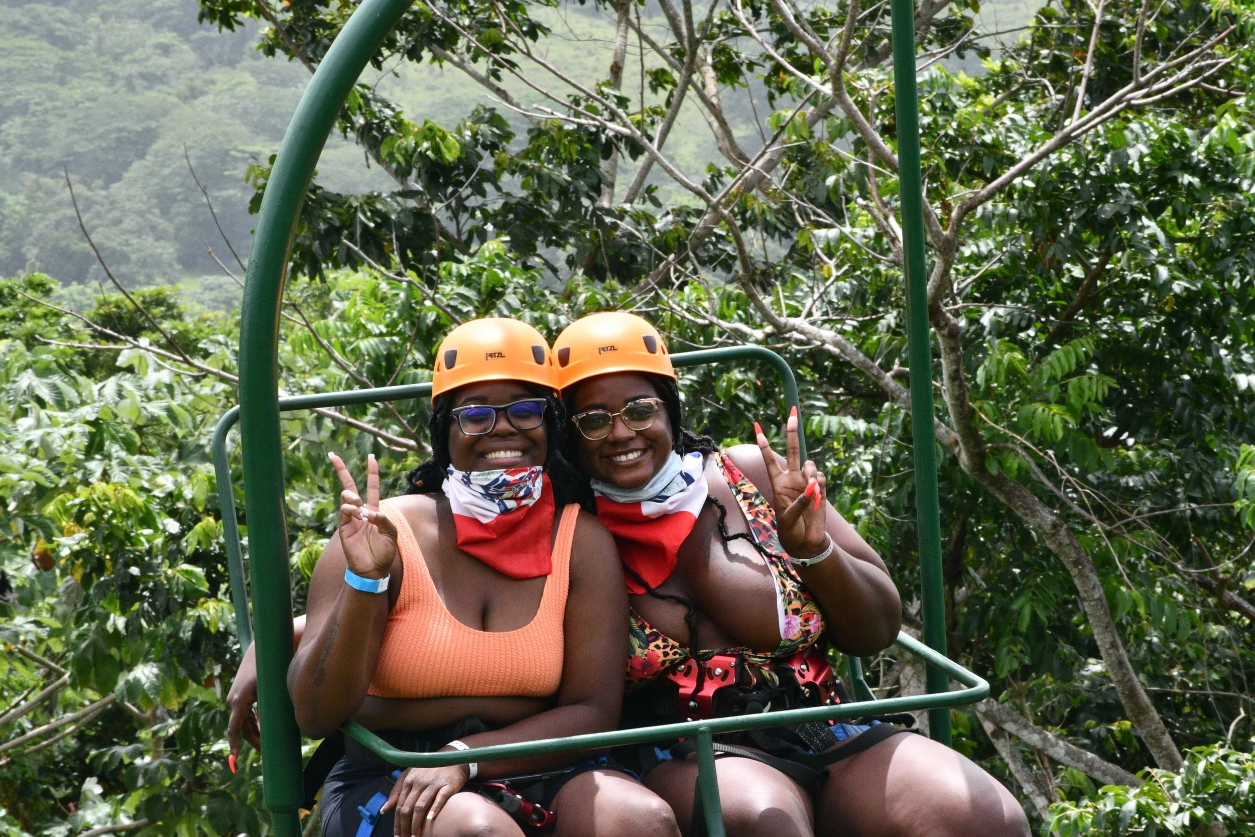 Two women wearing orange helmets and masks, making peace signs, sitting on a green cable car with a forest background.