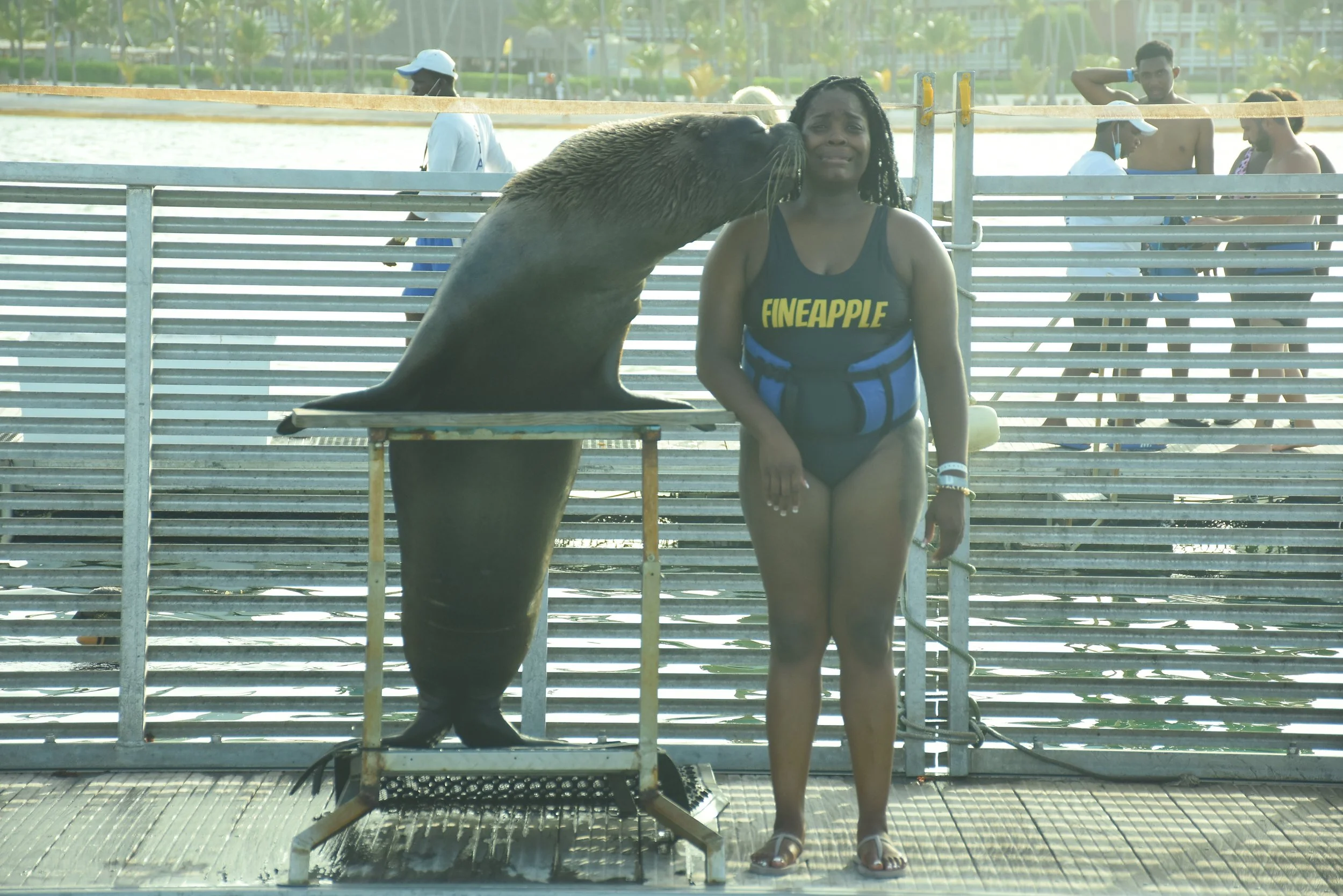 A woman standing next to a seal that is licking her face at a water park or marine zoo.