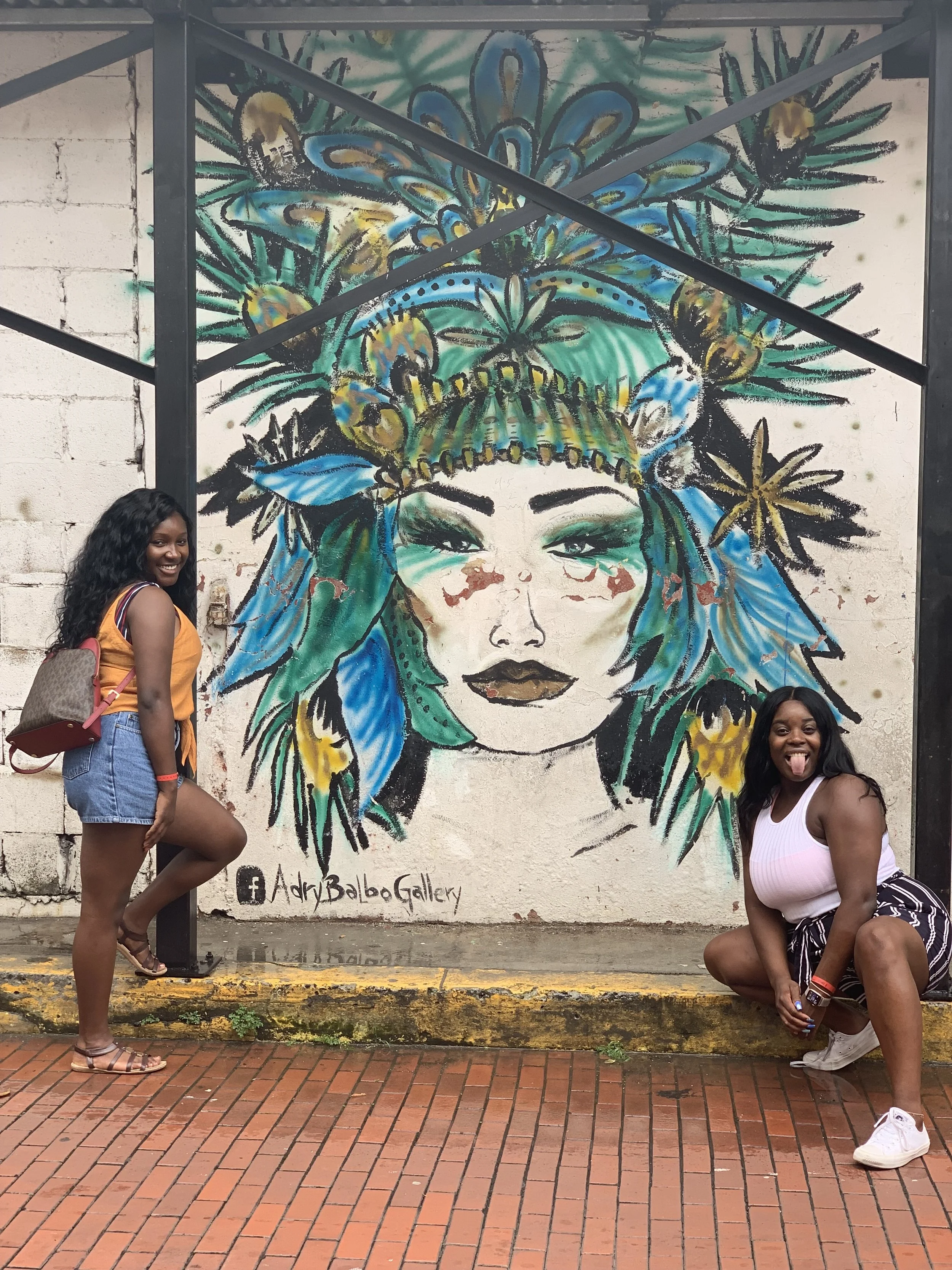 Two women posing in front of a large colorful mural of a woman's face with turquoise, blue, yellow, and green hair and floral headpiece. One woman stands on the left, smiling with a backpack, while the other squats on the right, sticking out her tong