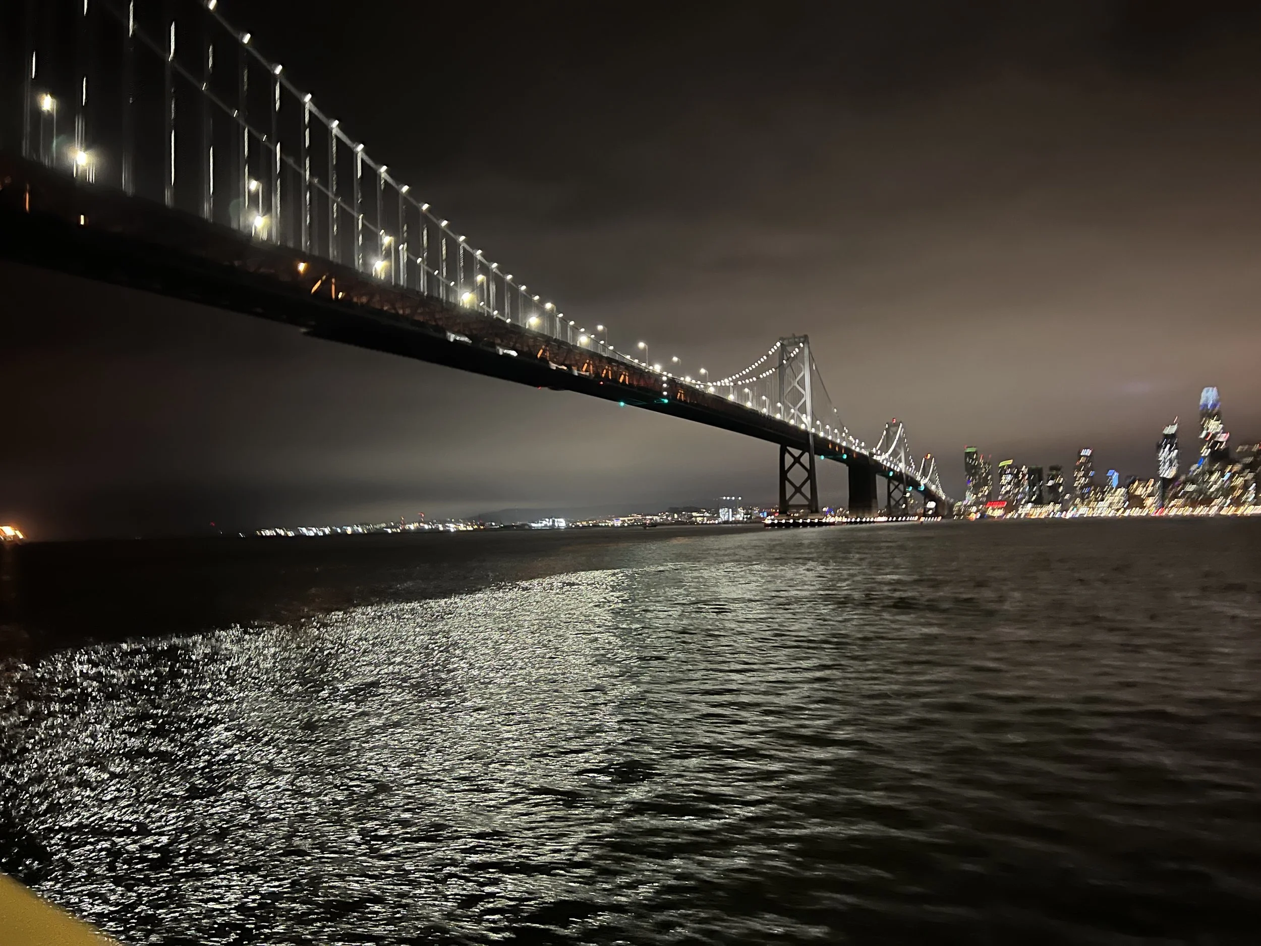 Nighttime view of a large illuminated suspension bridge over water with city skyline in the background.