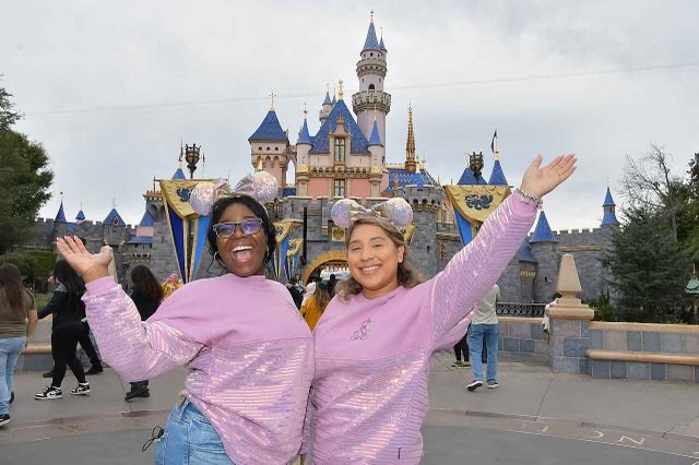 Two women smiling and celebrating in front of Sleeping Beauty Castle at Disneyland, wearing matching pink outfits and Mickey Mouse ears.