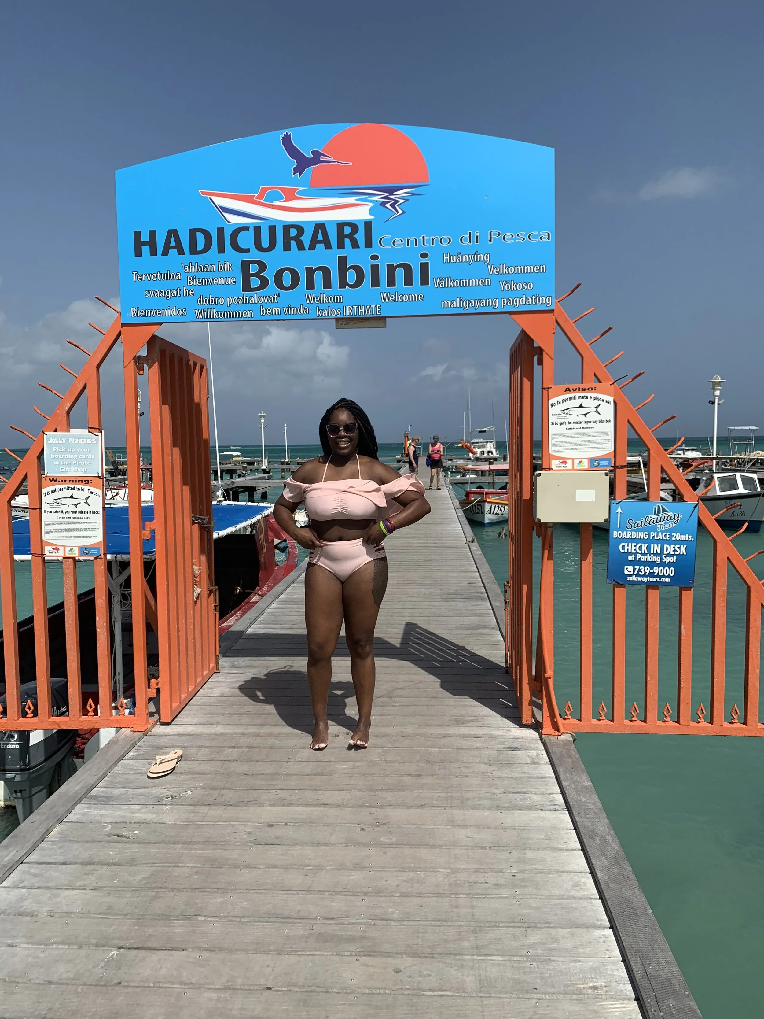 A woman in a pink bikini standing on a wooden pier at Hadicurari Bonbini fishing center in Aruba, with boats docked in the water and a cloudy sky in the background.