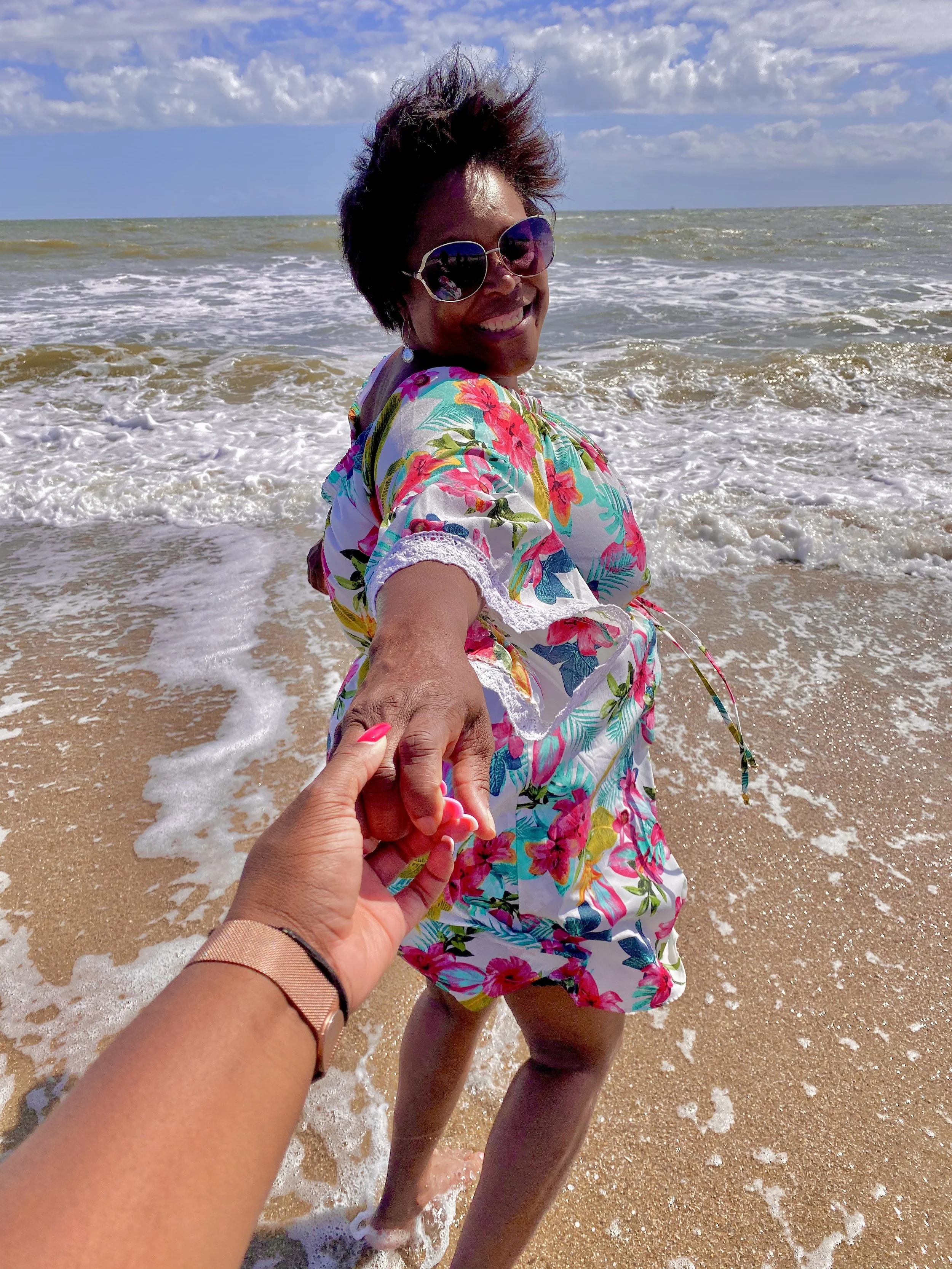 Smiling woman in sunglasses and a floral dress holding someone's hand at the beach with waves and blue sky in the background.