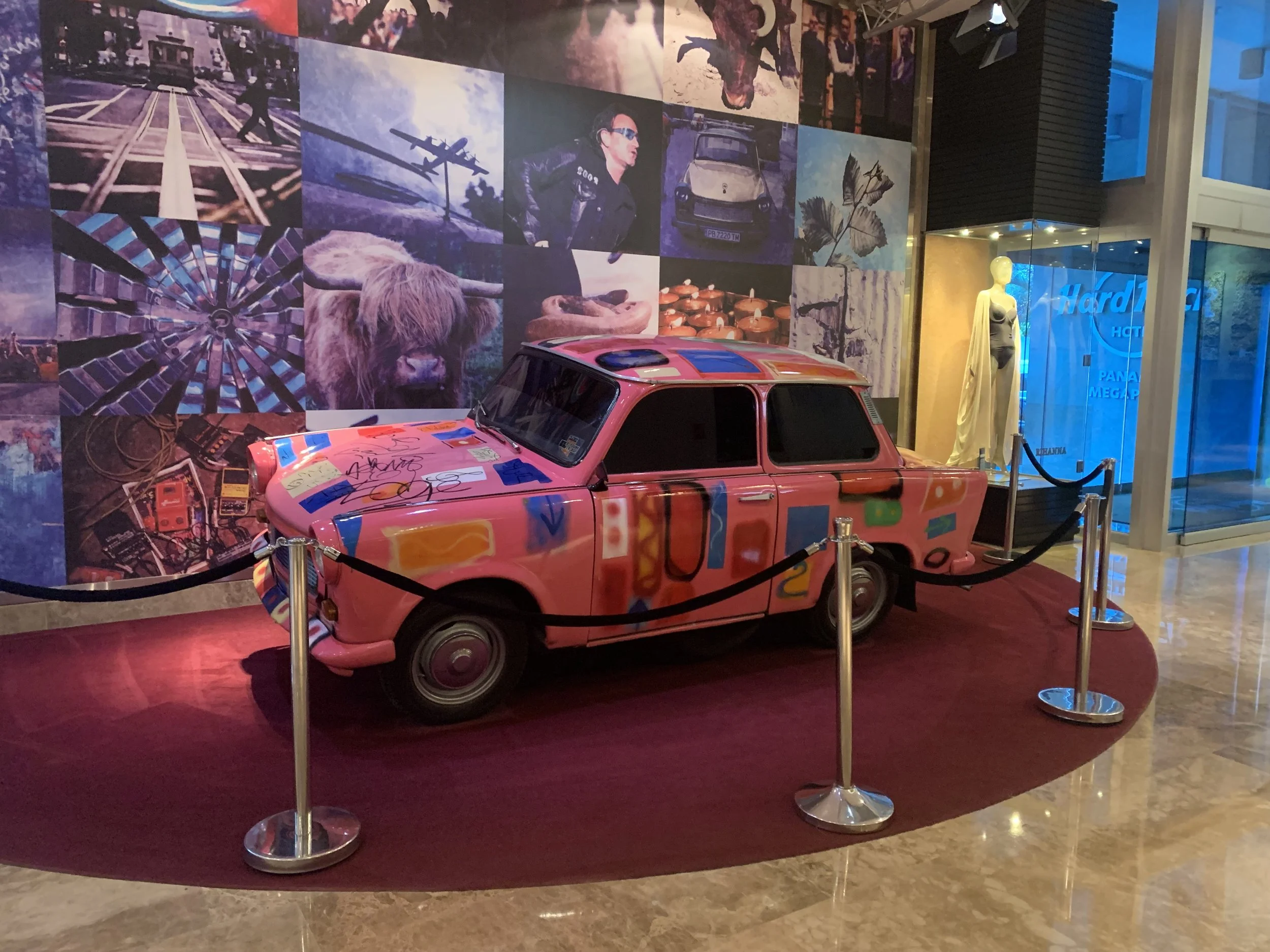 Pink vintage car with colorful graffiti-style decorations on display inside a hotel lobby, surrounded by a black rope barrier, with a collage of various images and a mannequin in a dress in the background.