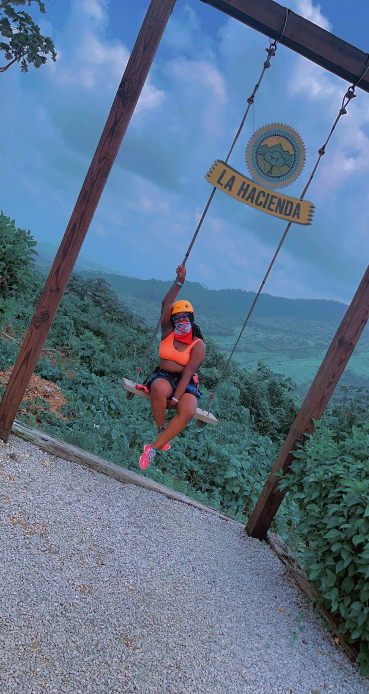 A woman wearing an orange helmet, a face mask, a bright orange top, black shorts, and pink shoes is sitting on a swing labeled 'La Hacienda' with a scenic landscape of green hills and blue sky in the background.