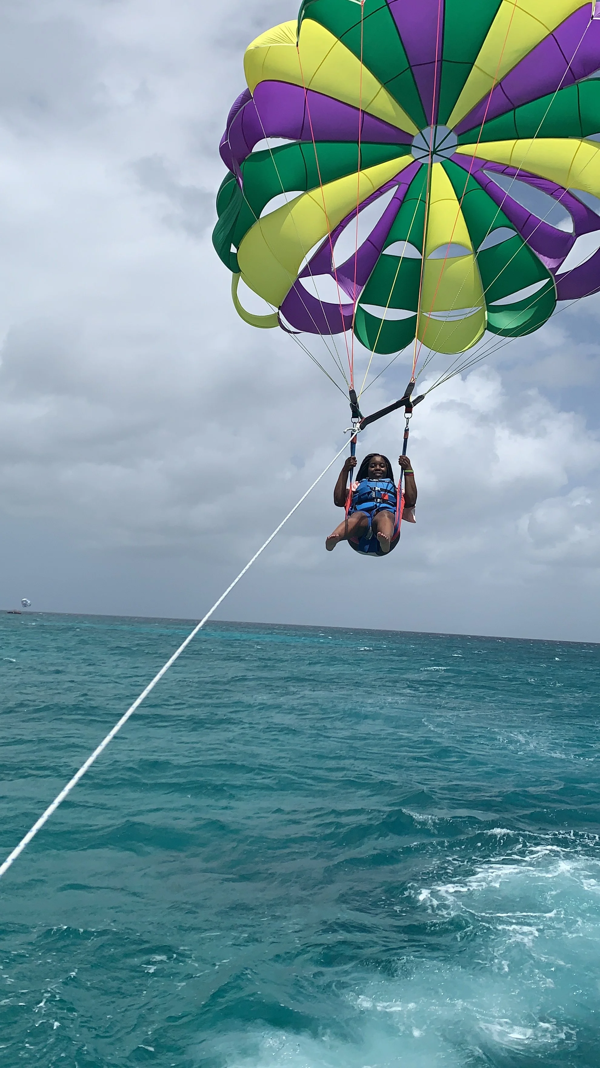 A person parasailing over the ocean under a colorful parachute with green, yellow, and purple sections, with cloudy sky in the background.