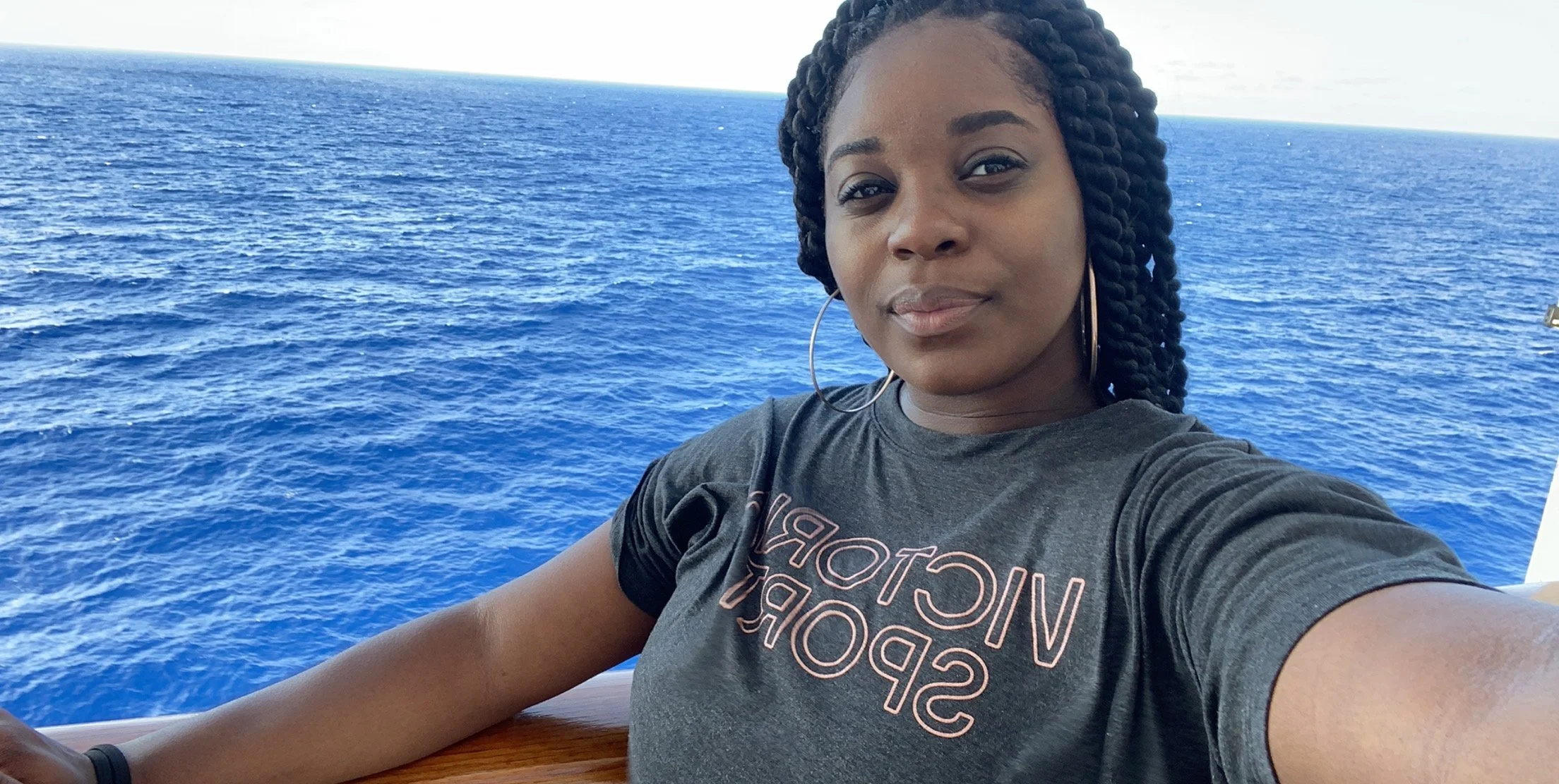 A woman taking a selfie in front of a vast blue ocean, wearing a dark t-shirt and hoop earrings.