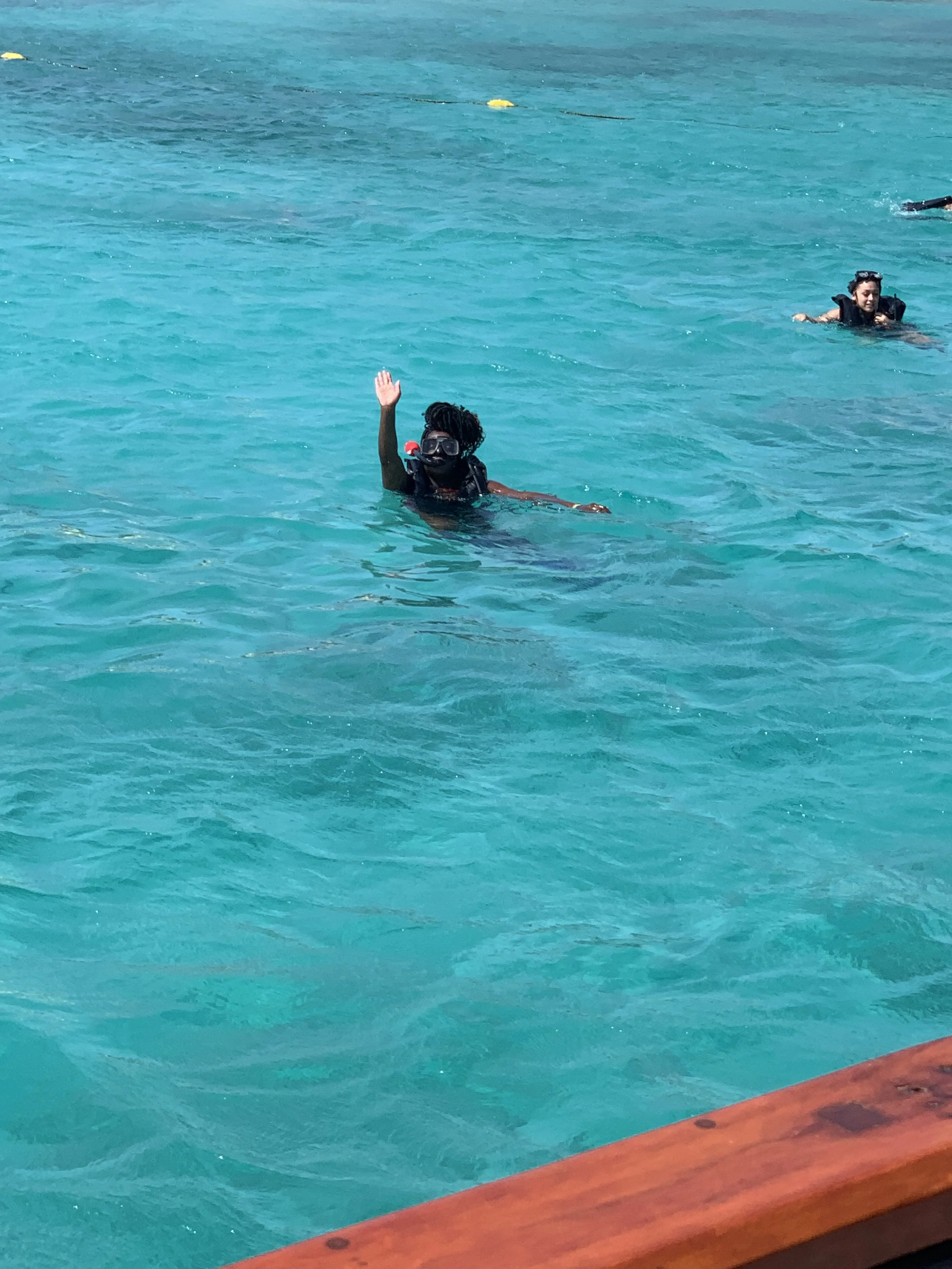 Person in the water wearing a diving mask and waving, with other people swimming in the background.