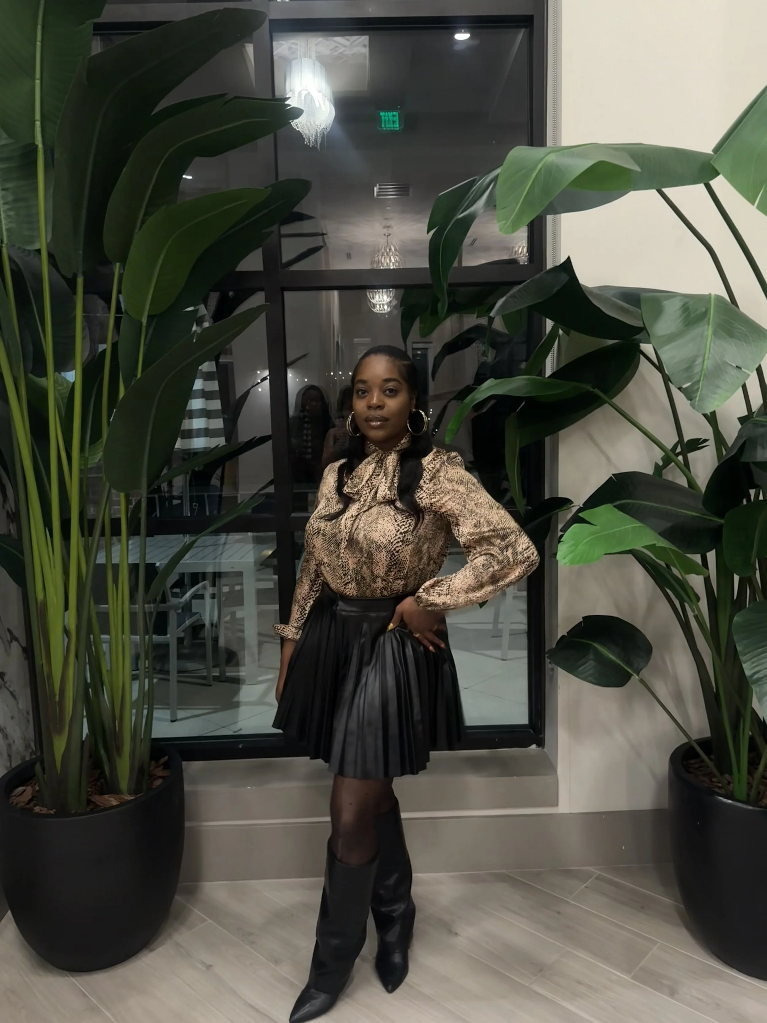 A woman standing in front of a glass window, surrounded by large green potted plants, wearing a snake print blouse, black pleated skirt, black over-the-knee boots, and hoop earrings.