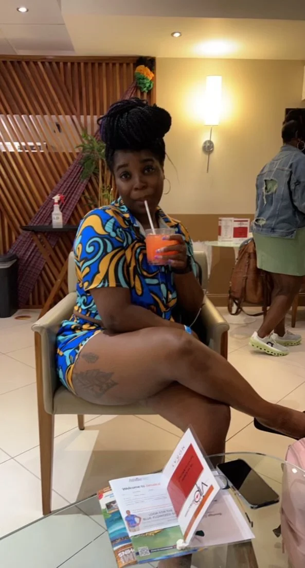 A woman sitting in a chair, drinking a pink beverage with a straw, wearing a colorful blue and yellow patterned outfit, and showcasing a tattoo on her thigh. She has her hair styled in a large braided bun.