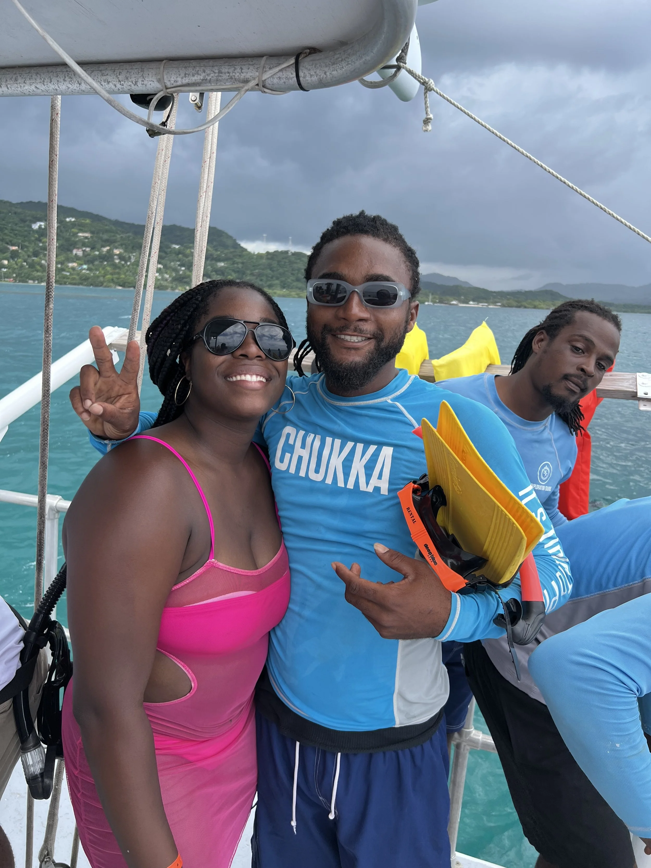 Group of three people on a boat with a scenic water and mountain background. Two women and one man, all with dark hair and medium to dark skin, wearing sunglasses and casual clothing. The woman in the pink dress is making a peace sign. The man in the