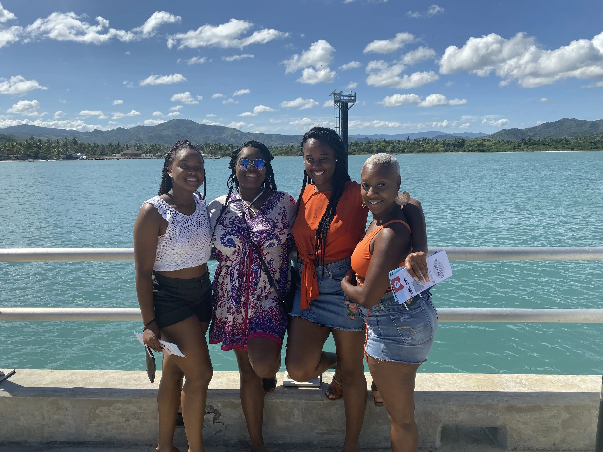 Four women standing together near a body of water with mountains and a cloudy sky in the background, smiling for the photo.
