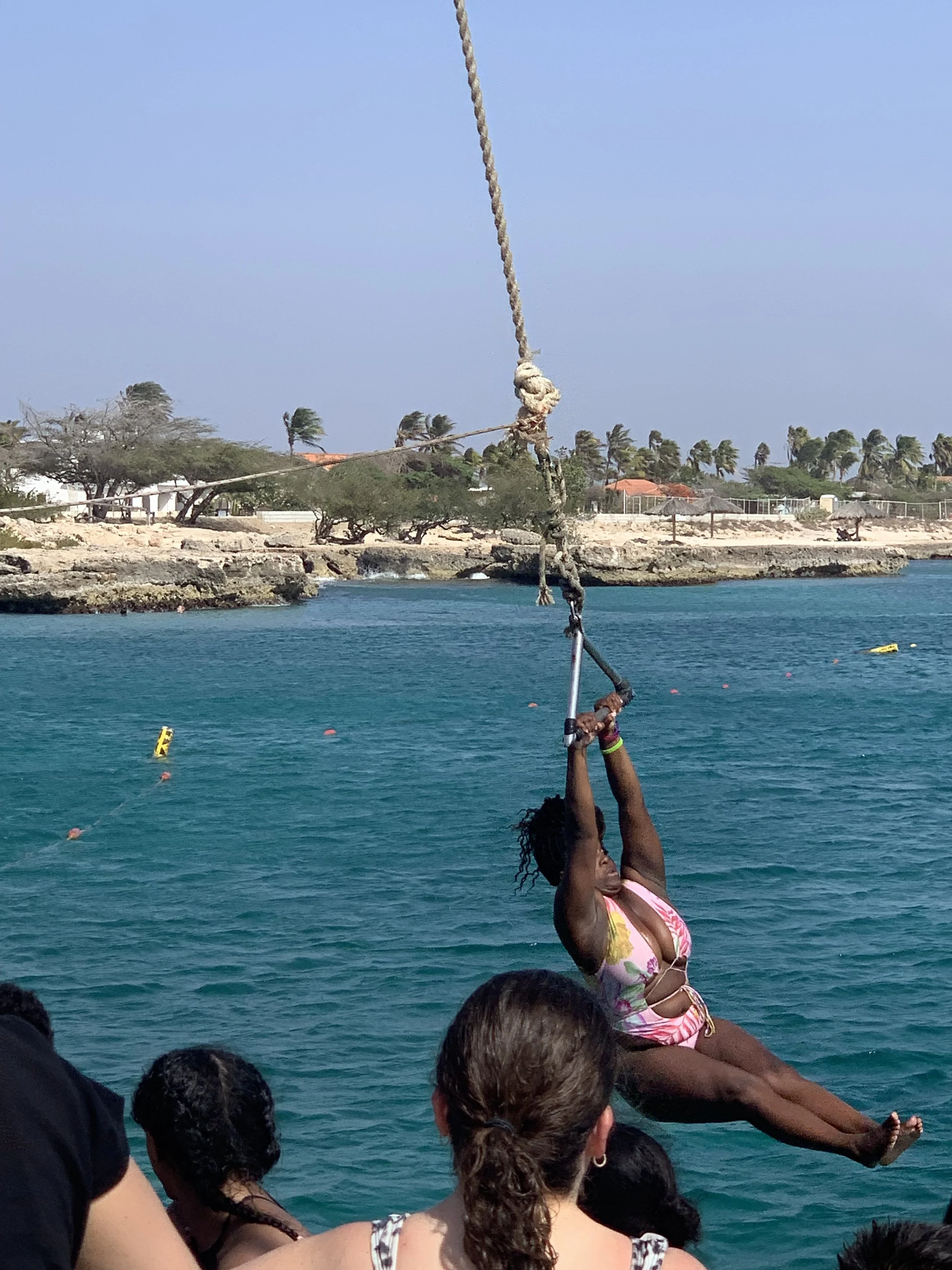 A woman in a pink swimsuit swinging on a rope over the water at a tropical beach, with onlookers watching.