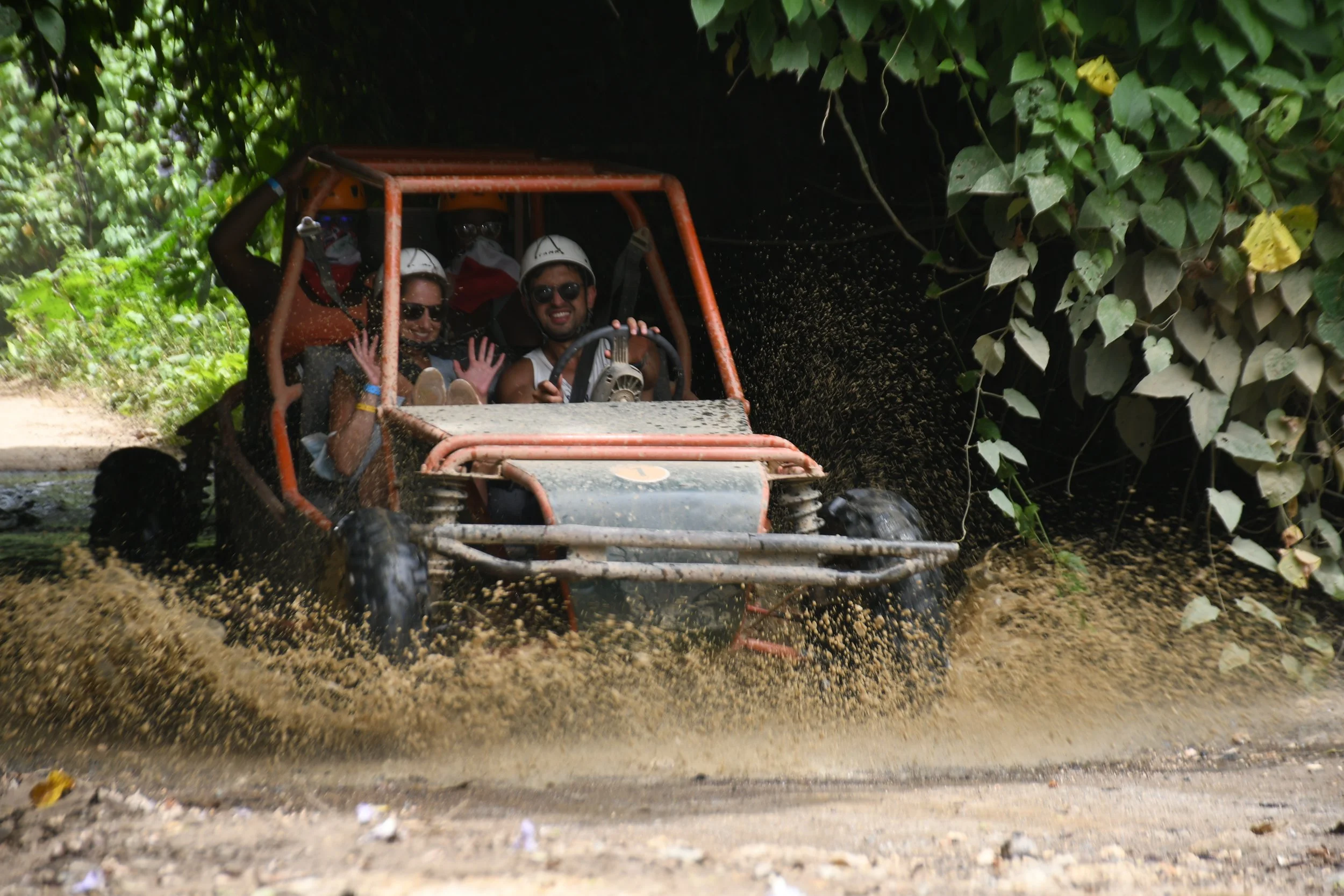 Group of friends enjoying off-road adventure in an open vehicle, driving through muddy terrain under a shady, leafy canopy, with water splashing around during the ride.