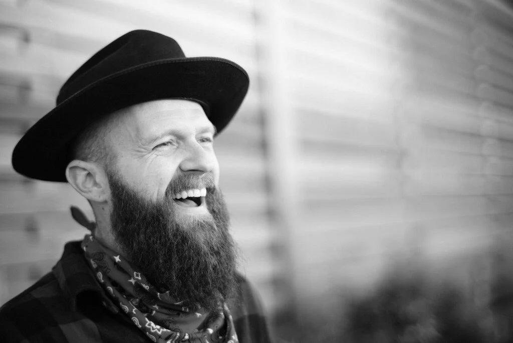 A smiling man with a long beard wearing a black hat and a bandana around his neck, standing against a blurred wooden background.
