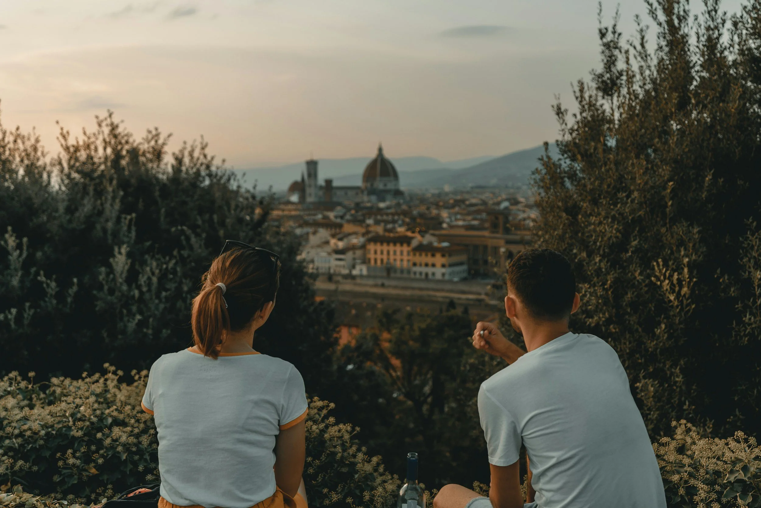 A man and woman sitting outdoors on a hillside, overlooking a view of Florence, Italy, with the Duomo in the background at sunset.