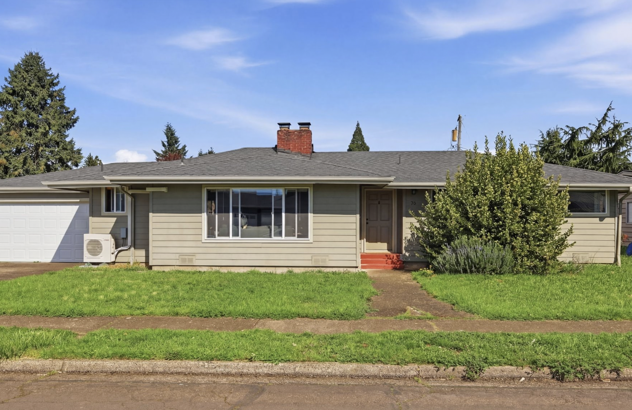 Front view of a single-story house with beige siding, a large front window, a small front porch with red steps, a white garage door, a large bush near the entrance, and a green lawn. There are trees in the background under a blue sky.