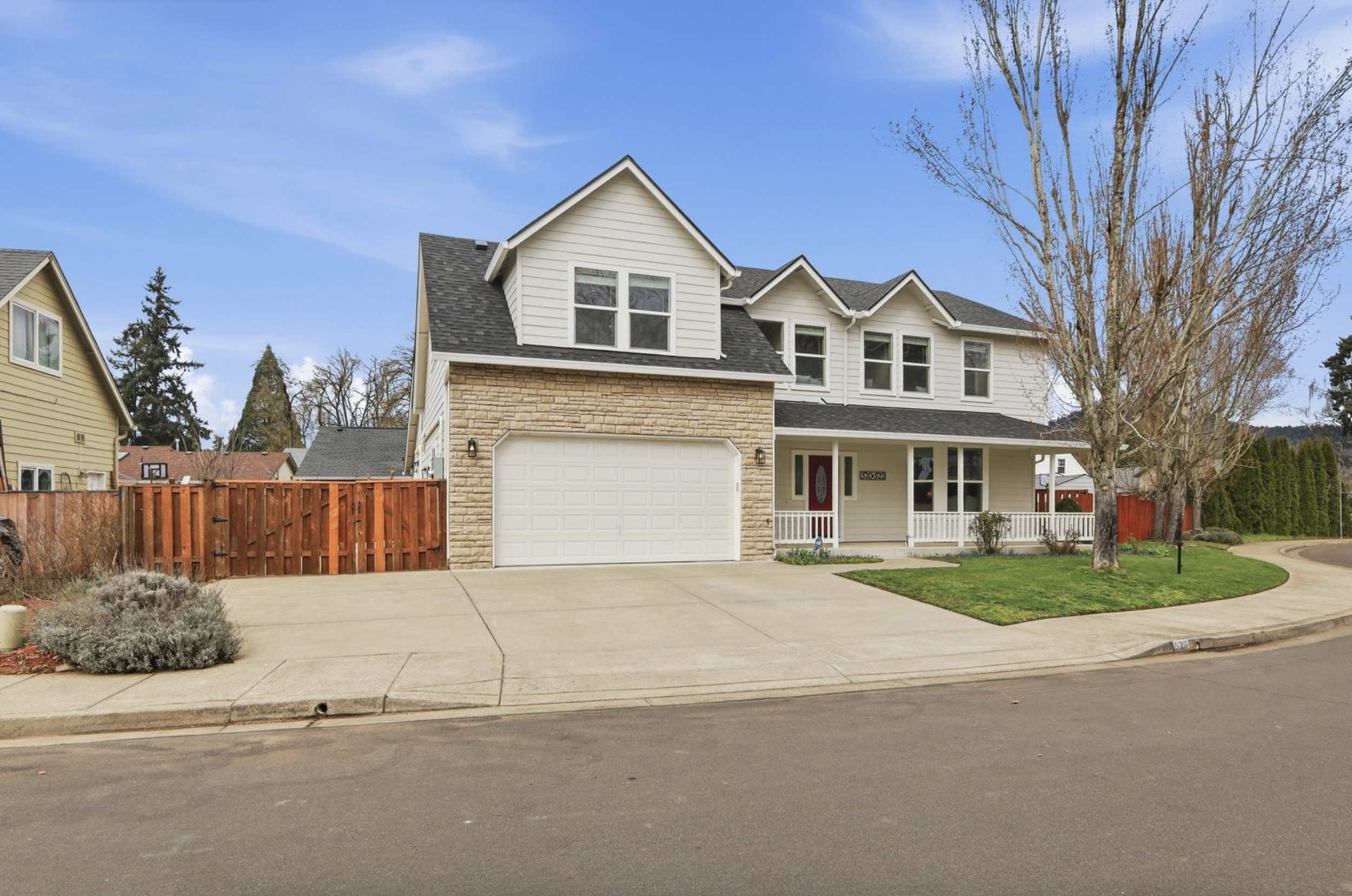 Front view of a two-story house with a garage, a small porch, a lawn, a tree, and a red wooden fence on the left side. Clear blue sky in the background.
