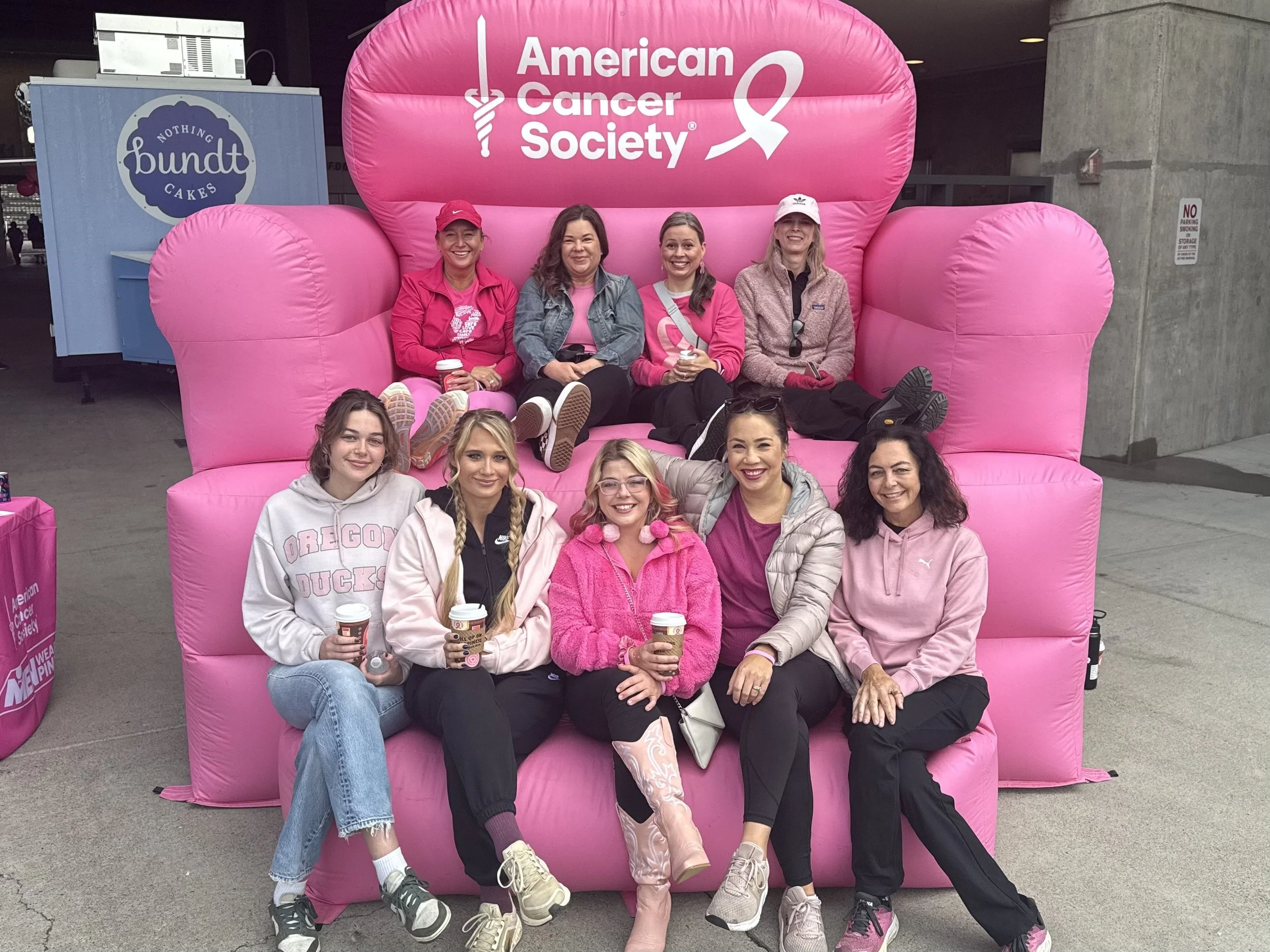 Group of women sitting and posing in front of a large pink inflatable structure with the American Cancer Society logo, most holding coffee cups.
