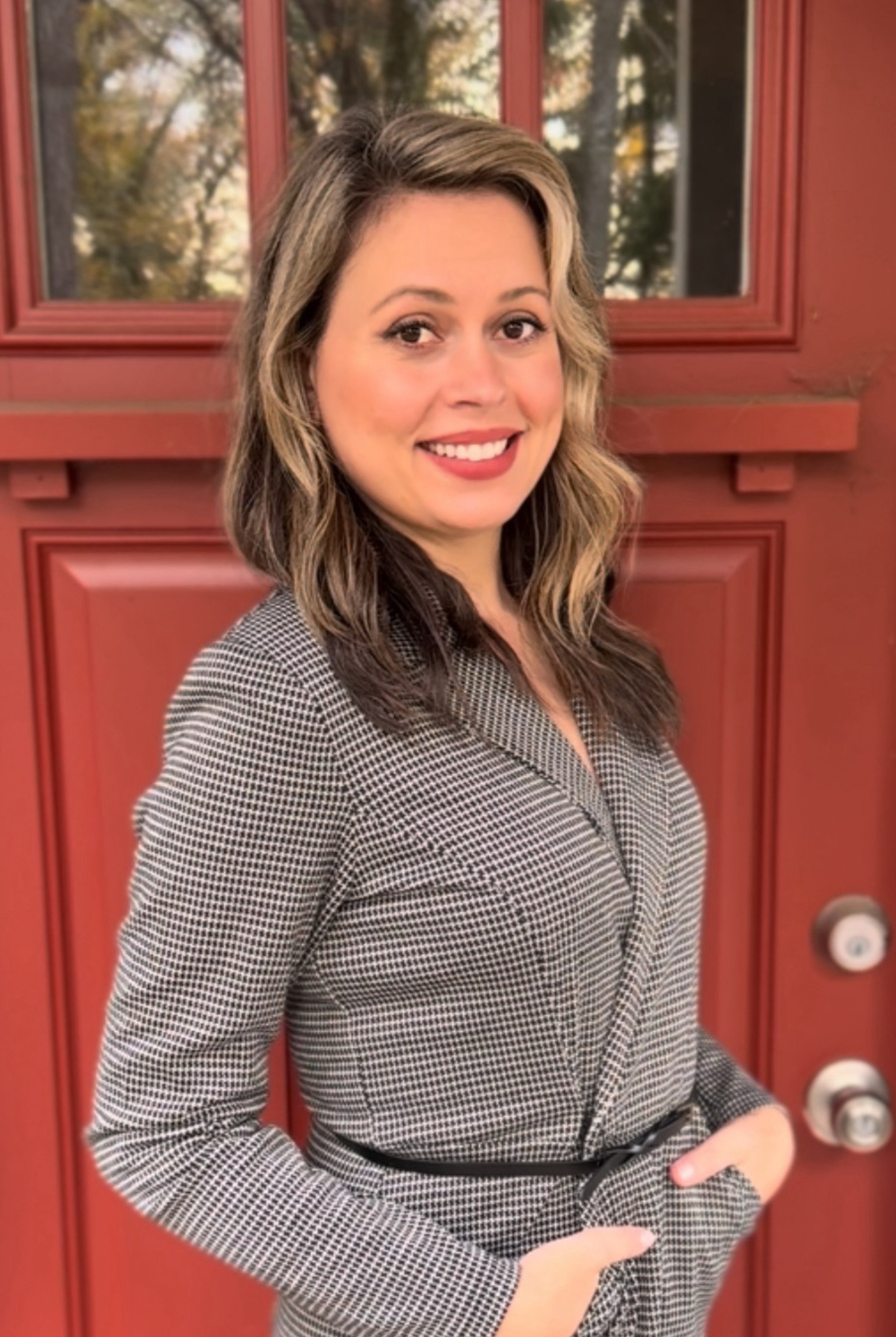 A woman with wavy, shoulder-length hair smiling, wearing a checkered blazer, standing in front of a red door with glass windows.