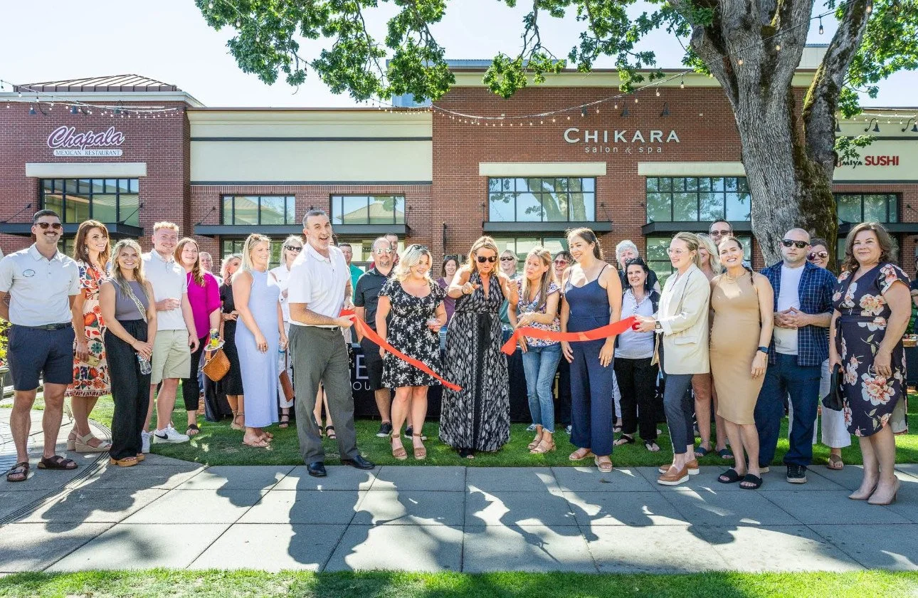 Group of people gathered outdoors for a ribbon-cutting ceremony in front of a shopping plaza with businesses like 'Chikara Salon & Spa' and 'Chapala Mexican Restaurant.'